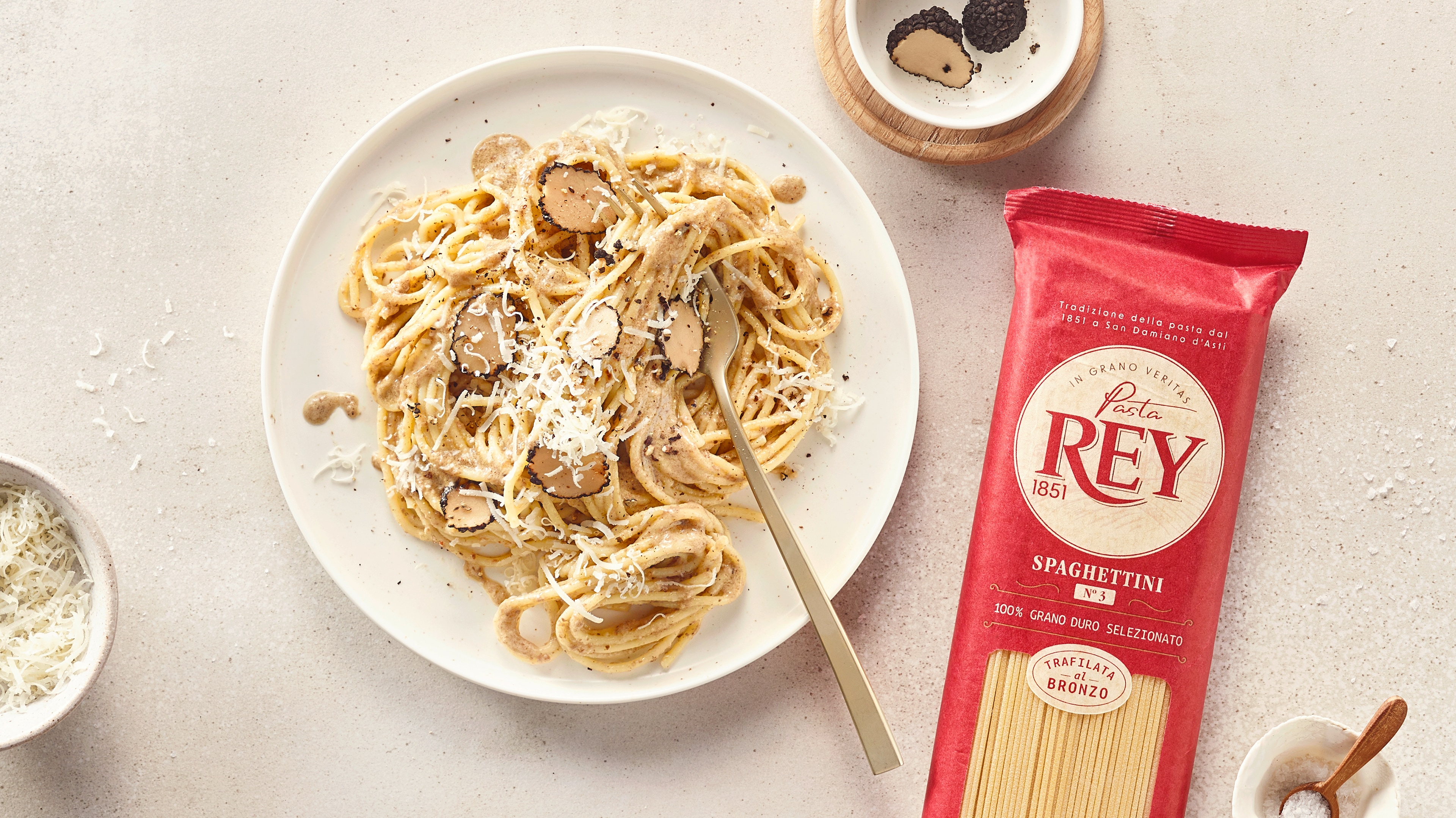 Teller mit Spaghetti, Trüffelscheiben und geriebenem Käse. Daneben eine Packung Pasta Rey Spaghettini