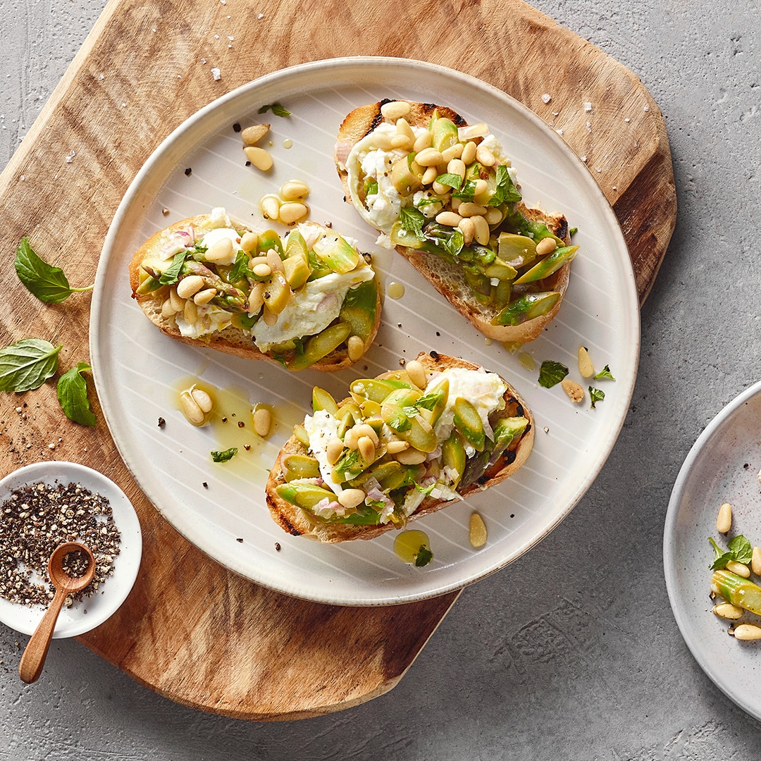 Ein Teller mit drei gerösteten Brotscheiben, darauf weiße Creme, grüne Spargelstücke, gehackte Kräuter und hellen Pinienkernen.