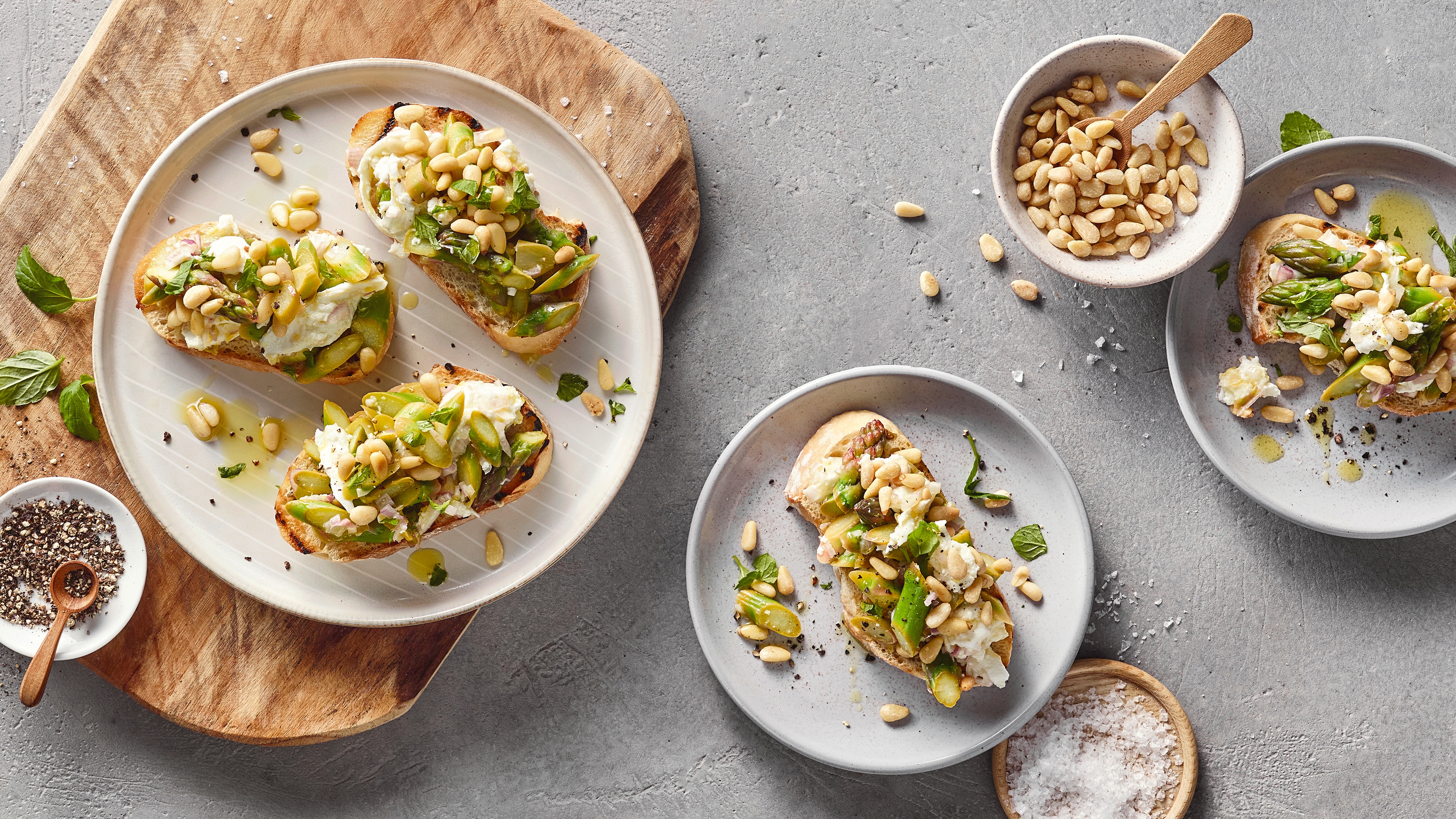 Ein Teller mit drei gerösteten Brotscheiben, darauf weiße Creme, grüne Spargelstücke, gehackte Kräuter und hellen Pinienkernen.