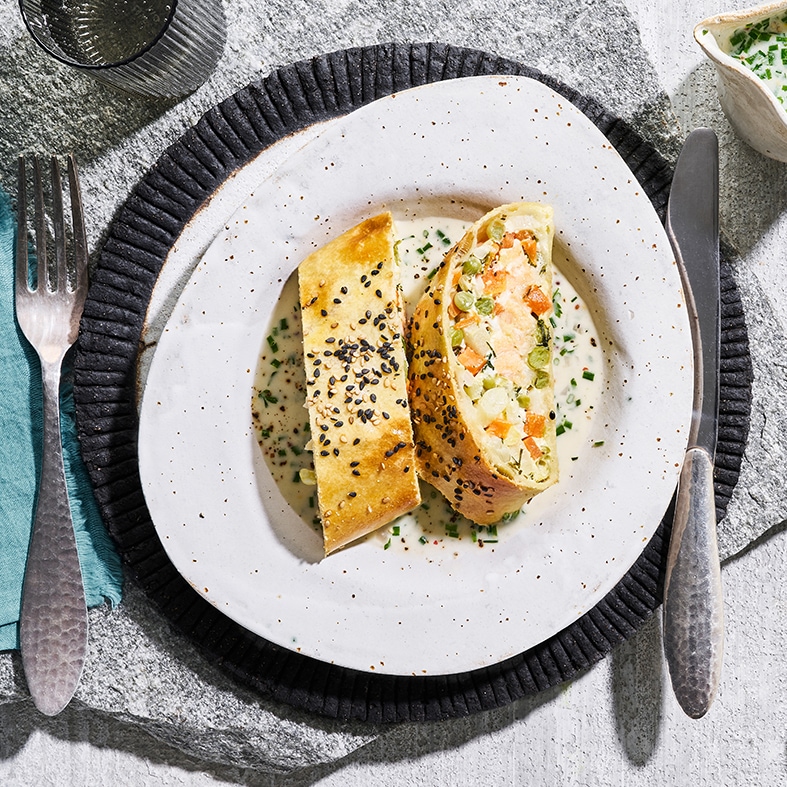 Unregelmäßig runder Teller mit zwei Lachs‑Gemüse‑Strudeln, bestreut mit Sesam; links eine Gabel und ein Glas Wasser, rechts ein Messer und etwas Petersilien-Dip.