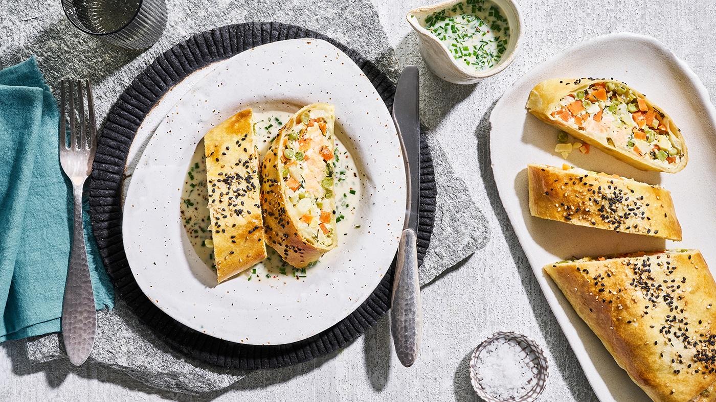 Unregelmäßig runder Teller mit zwei Lachs‑Gemüse‑Strudeln, bestreut mit Sesam; links eine Gabel und ein Glas Wasser, rechts ein Messer und etwas Petersilien-Dip. Rechts liegt ein ganzer angeschnittener Strudel.