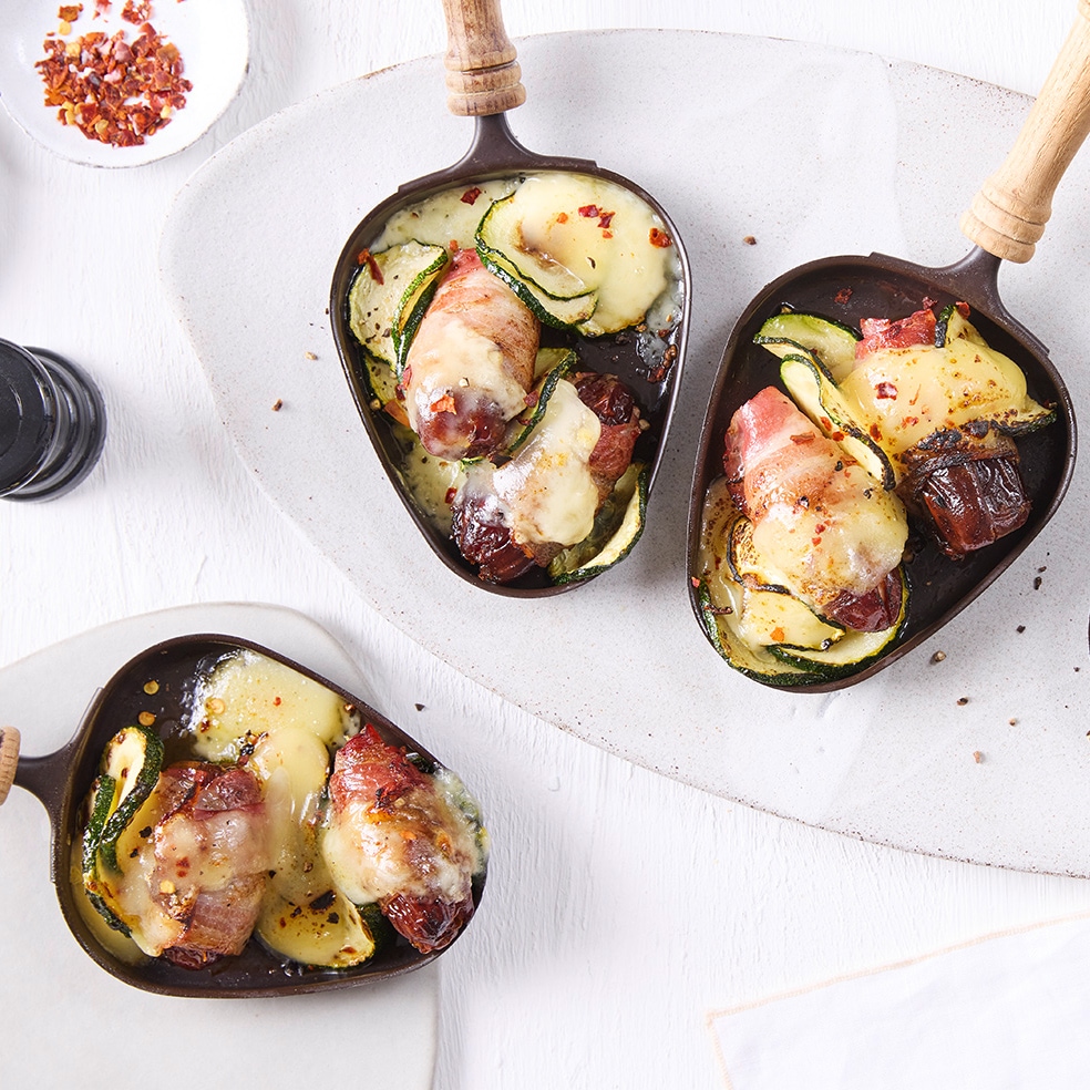 Gusseisenpfännchen mit geschmolzenem Käse, Speckdatteln, Zucchinischeiben und roten Chiliflocken