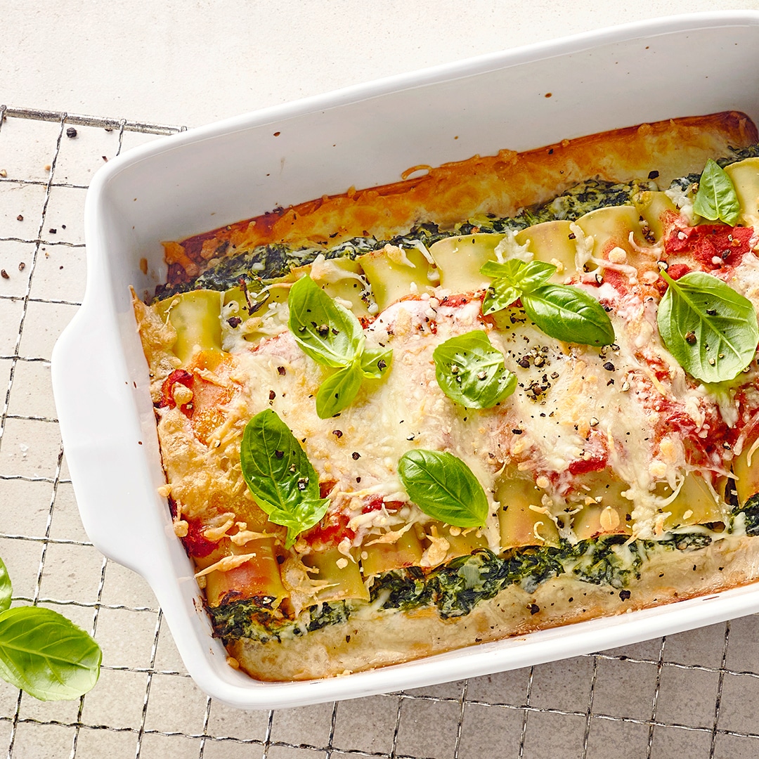 Eine Auflaufform mit Nudelauflauf aus Cannelloni, Spinat, Käse und Tomatensoße, obenauf liegen frische Basilikumblätter