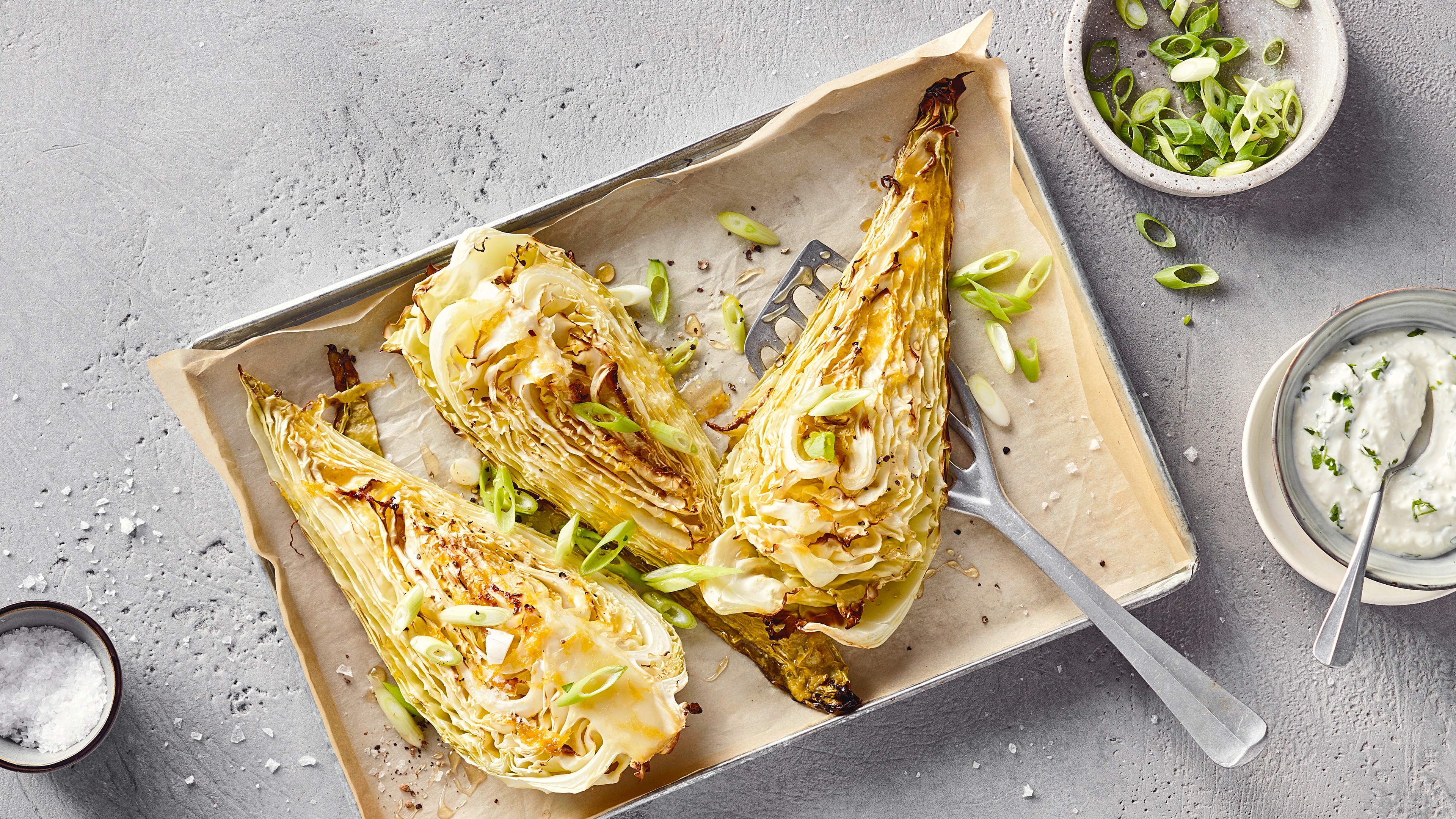 Rezept für Spitzkohl aus dem Ofen Ein Backblech mit halbiertem, gebackenem Spitzkohl, garniert mit grünen Frühlingszwiebeln, daneben helle Creme