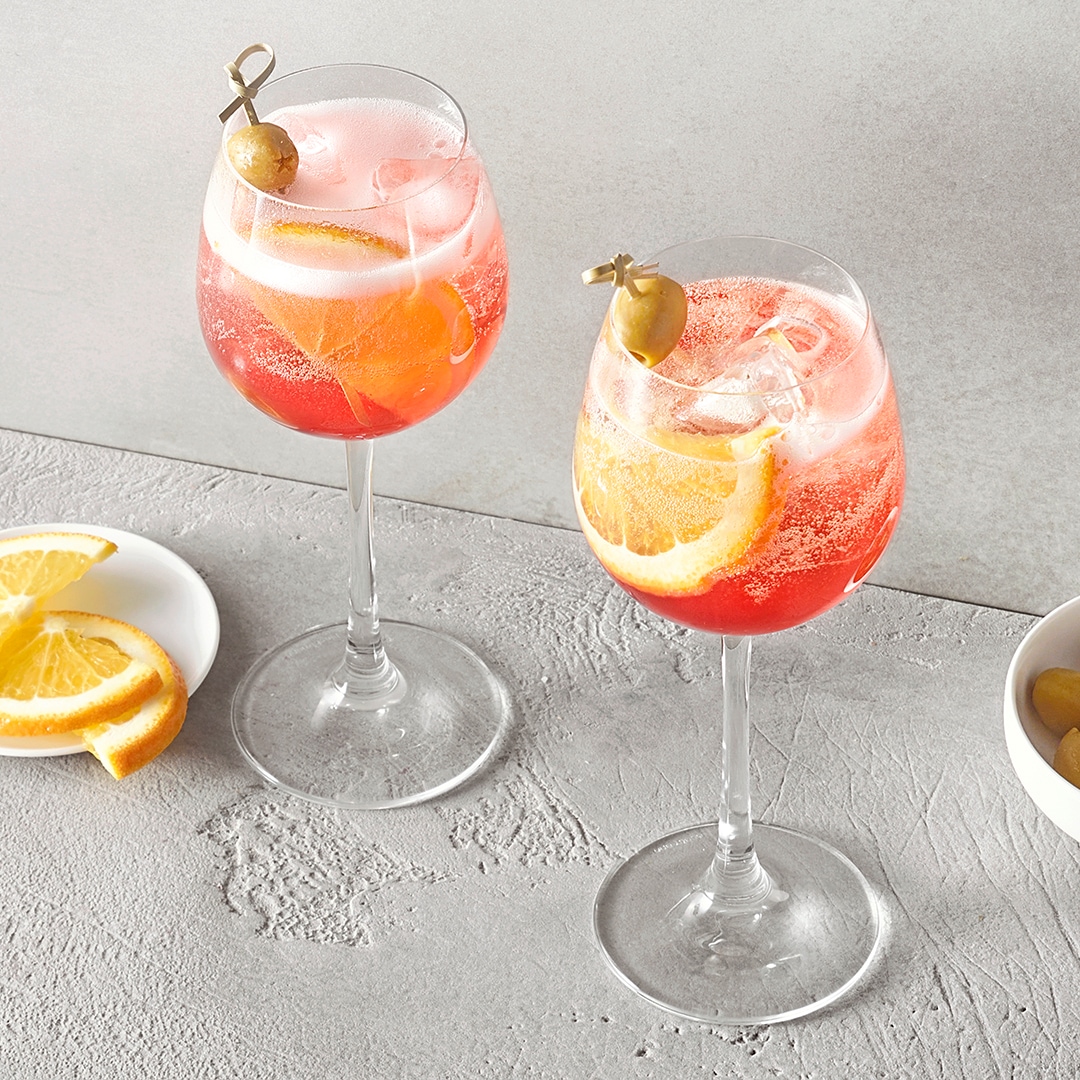 Ein Glas mit rosafarbenem Getränk, Eiswürfeln, Zitronenscheiben und einer grünen Olive am Spieß.