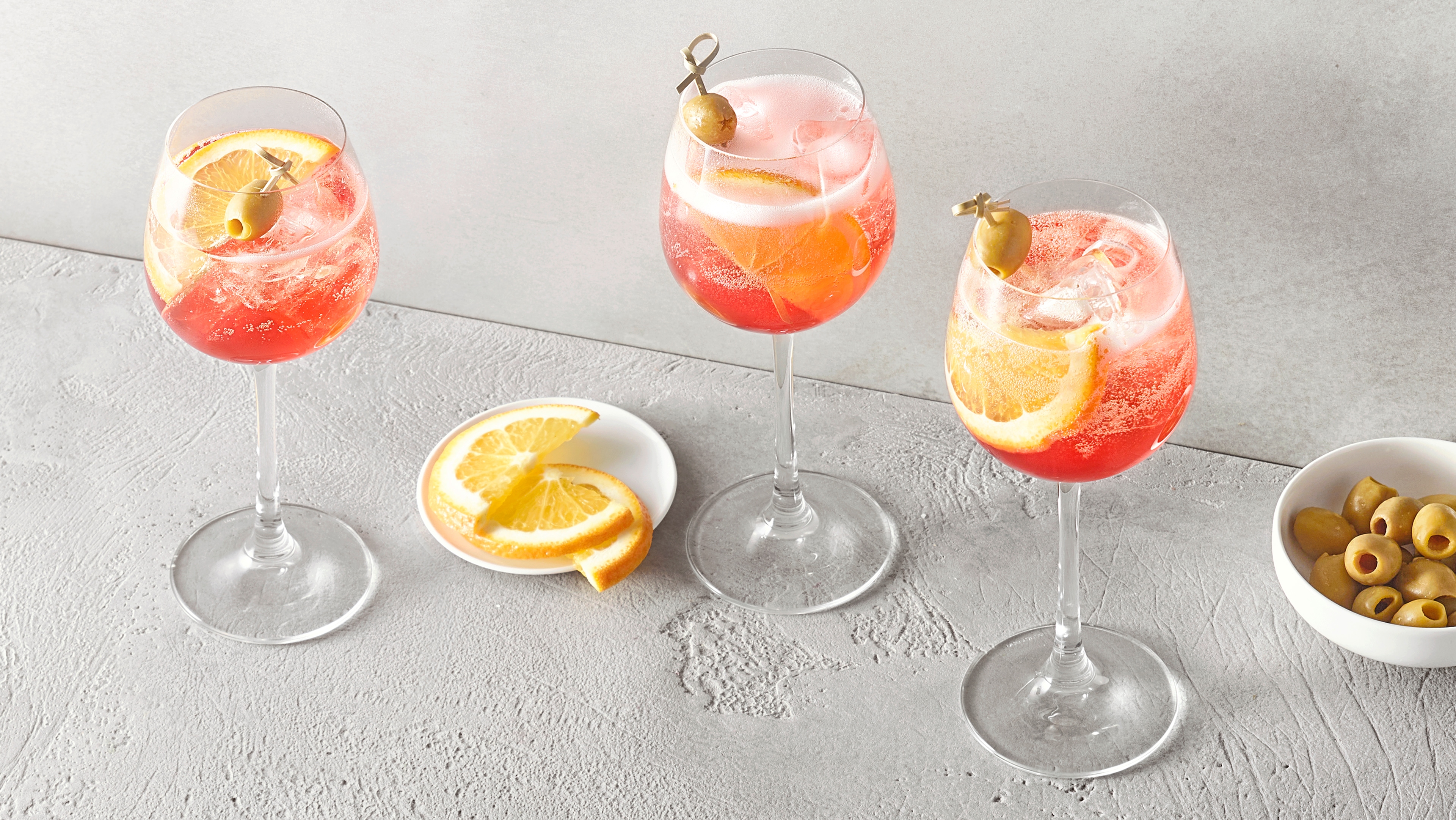 Ein Glas mit rosafarbenem Getränk, Eiswürfeln, Zitronenscheiben und einer grünen Olive am Spieß.