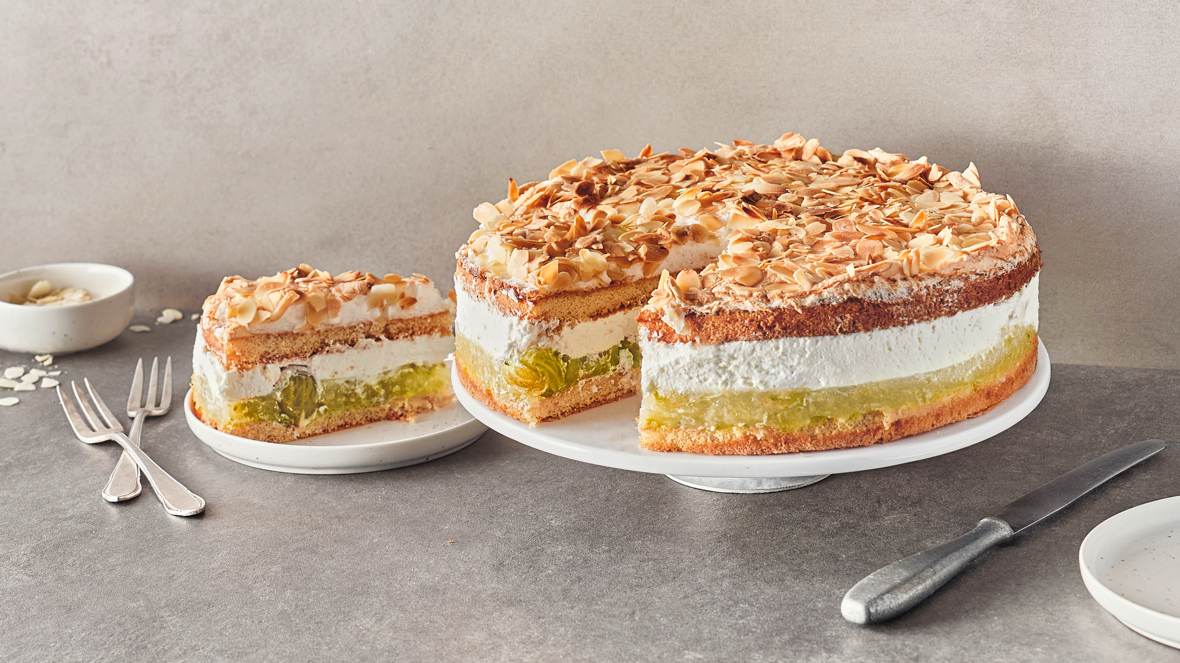 Eine Torte aus fluffigen hellen Böden, luftiger Creme und einer Fruchtschicht, getoppt mit Baiser und gehobelten Mandeln