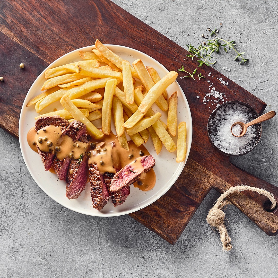 Ein Teller mit saftigen Fleischstücken, in der Mitte leicht rosa, getoppt mit heller Soße mit Pfefferkörnern, dazu knusprige Pommes