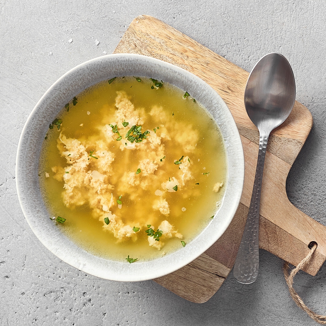 Eine Schüssel mit klarer gelber Brühe, darin helle lockere Klößchen und grün gehackte Kräuter.