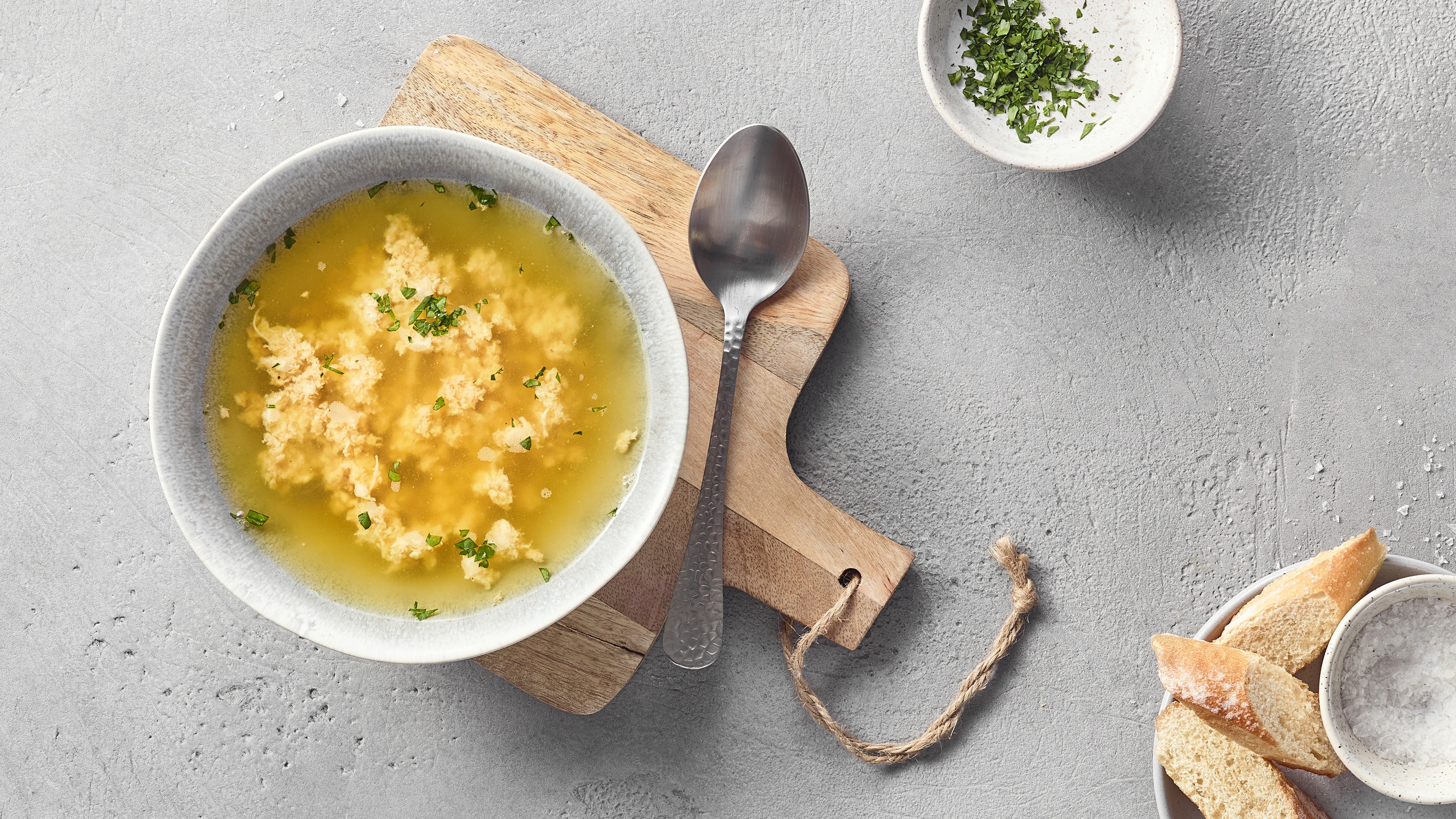 Eine Schüssel mit klarer gelber Brühe, darin helle lockere Klößchen und grün gehackte Kräuter.