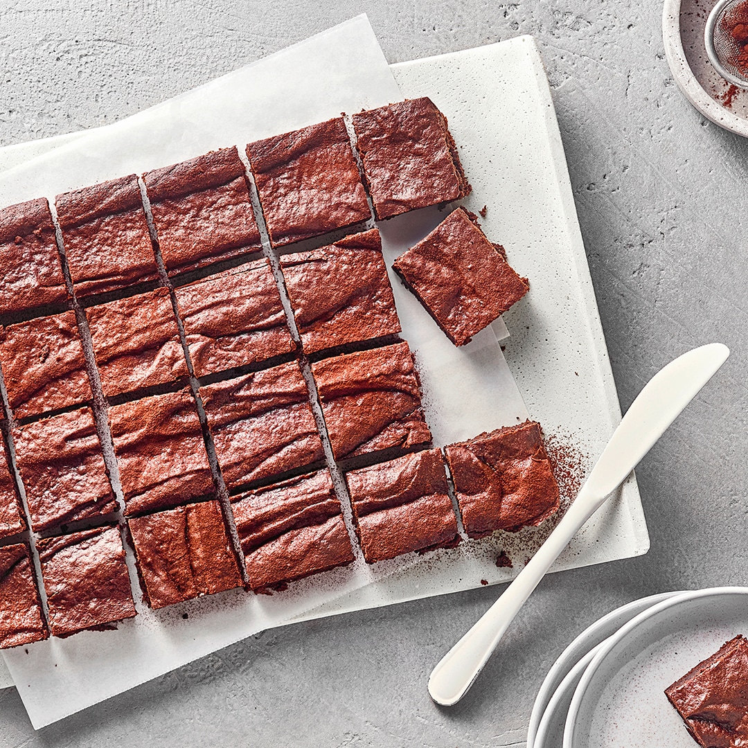 In Stücke geschnittene Schokoladen-Brownies auf einer Servierplatte mit Kakaopulver und Dessertteller im Hintergrund.