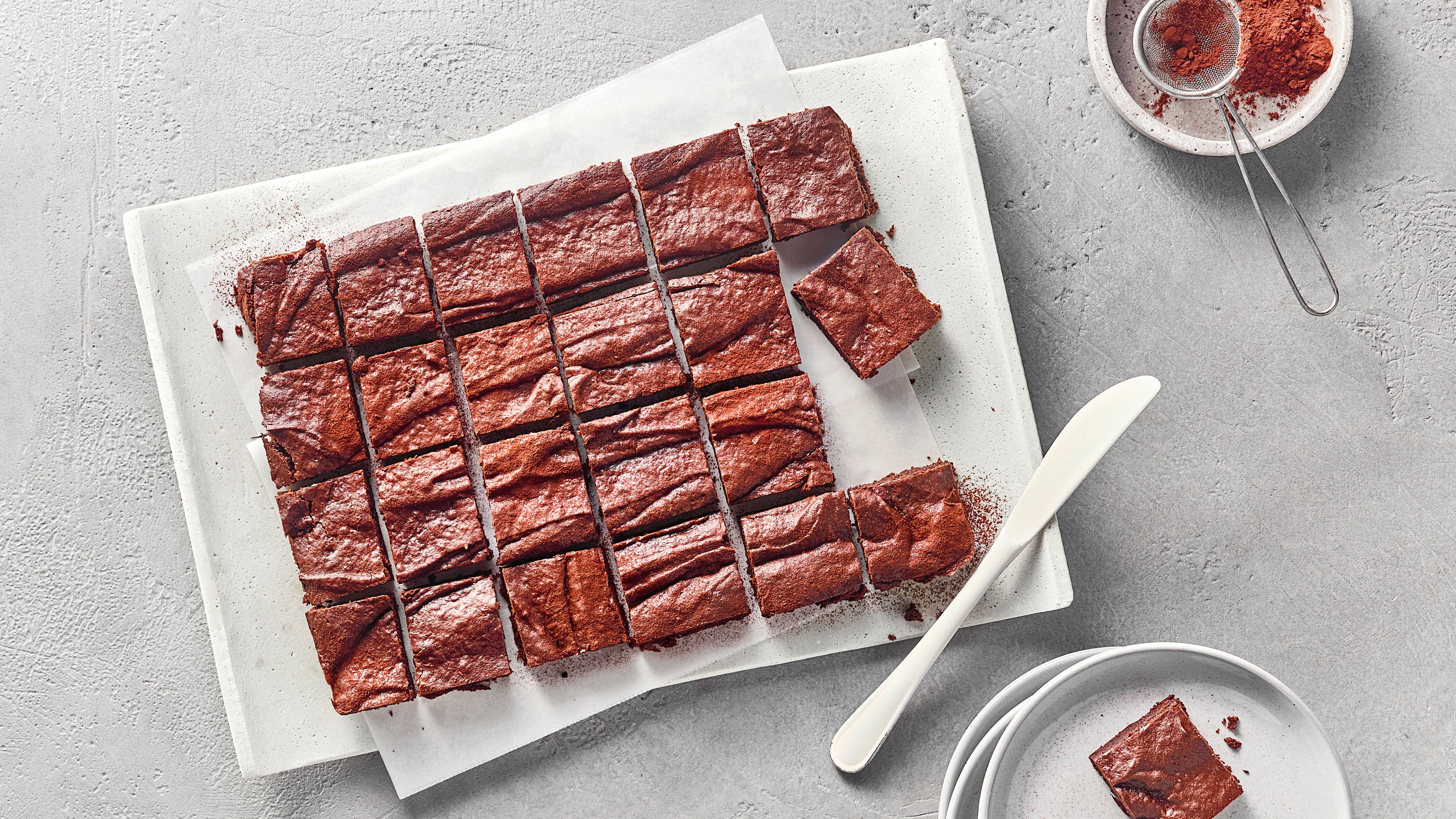 In Stücke geschnittene Schokoladen-Brownies auf einer Servierplatte mit Kakaopulver und Dessertteller im Hintergrund.