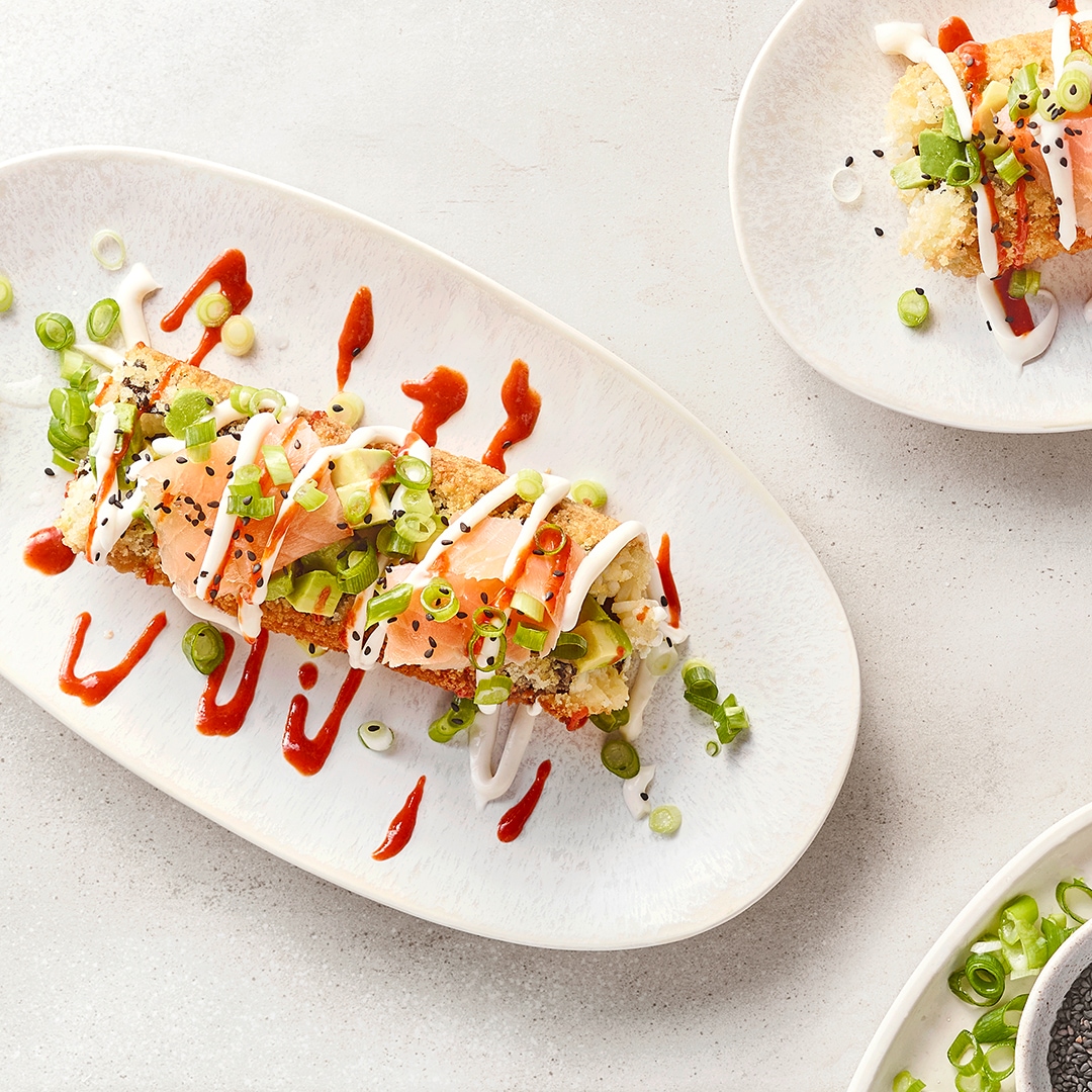 Zwei kunstvoll angerichtete Sushi-Rollen auf weißen Tellern, garniert mit Frühlingszwiebeln, Sauce und schwarzem Sesam.