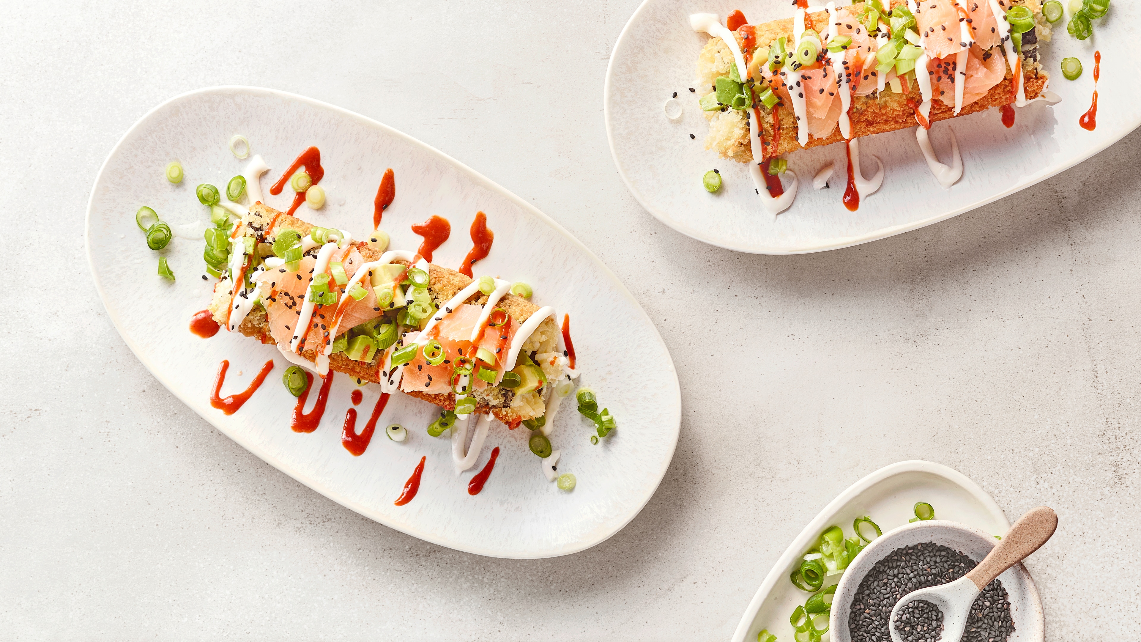 Zwei kunstvoll angerichtete Sushi-Rollen auf weißen Tellern, garniert mit Frühlingszwiebeln, Sauce und schwarzem Sesam.