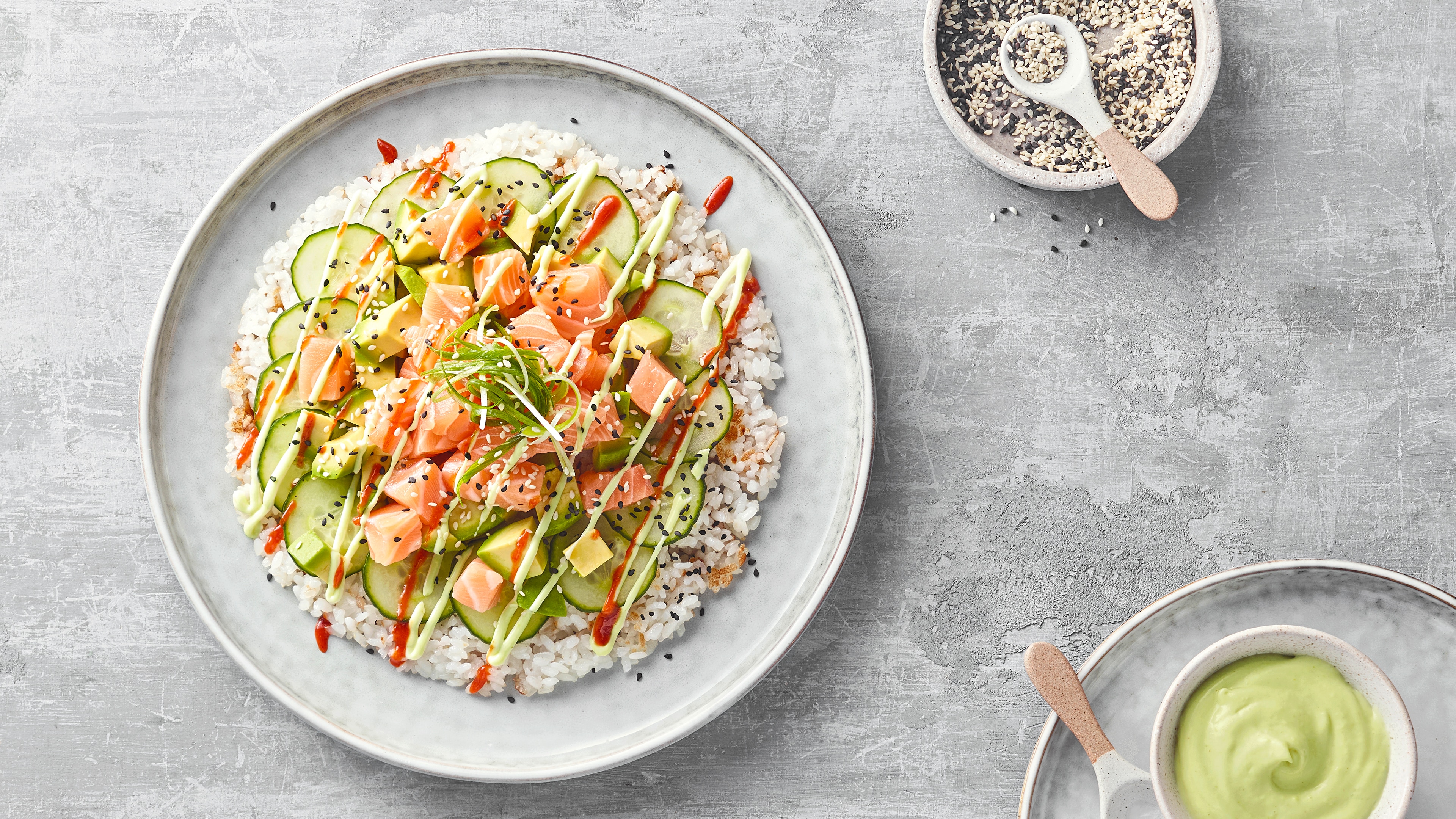Teller mit in Kreisform gedrückten Reis, Lachs, Avocado und Gurke, garniert mit Sauce und Sesam.