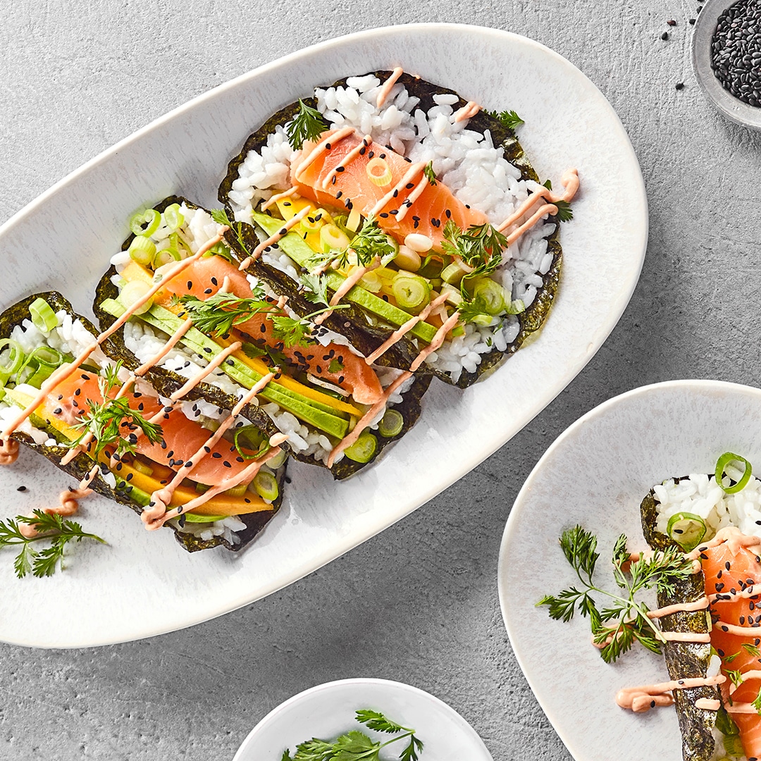 Sushi Tacos mit Reis, Lachs, Frühlingszwiebeln und Sauce auf Nori-Blättern, angerichtet auf weißem Teller.