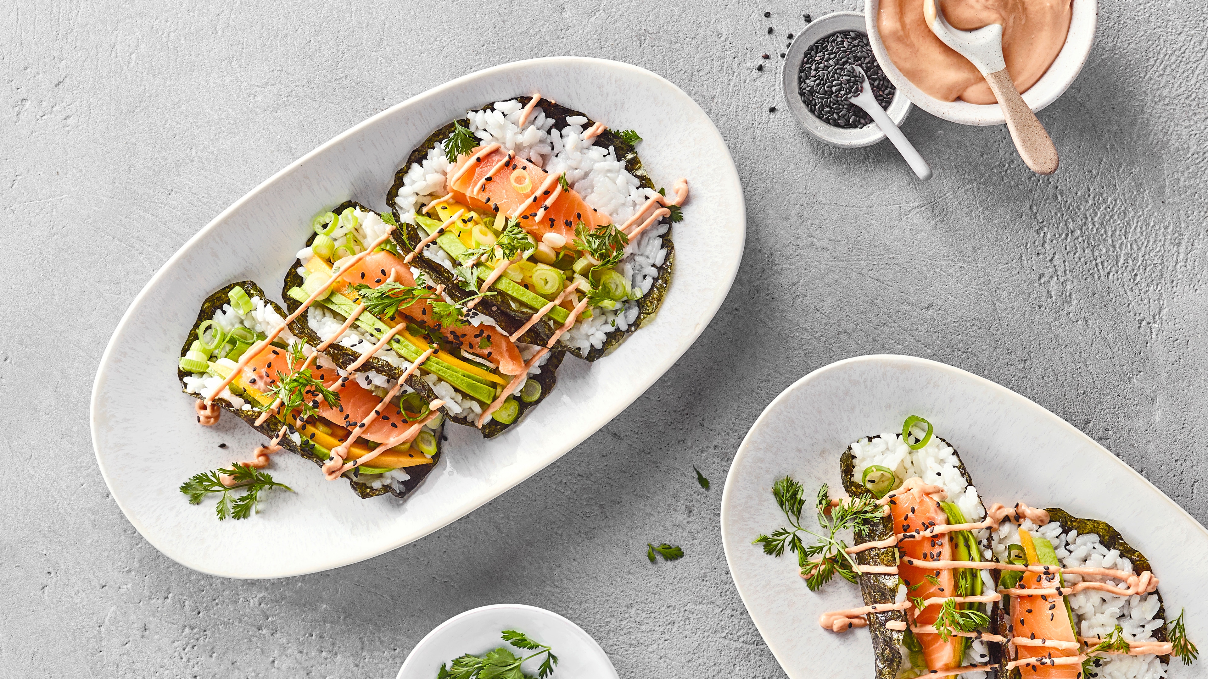 Sushi Tacos mit Reis, Lachs, Frühlingszwiebeln und Sauce auf Nori-Blättern, angerichtet auf weißen Tellern.