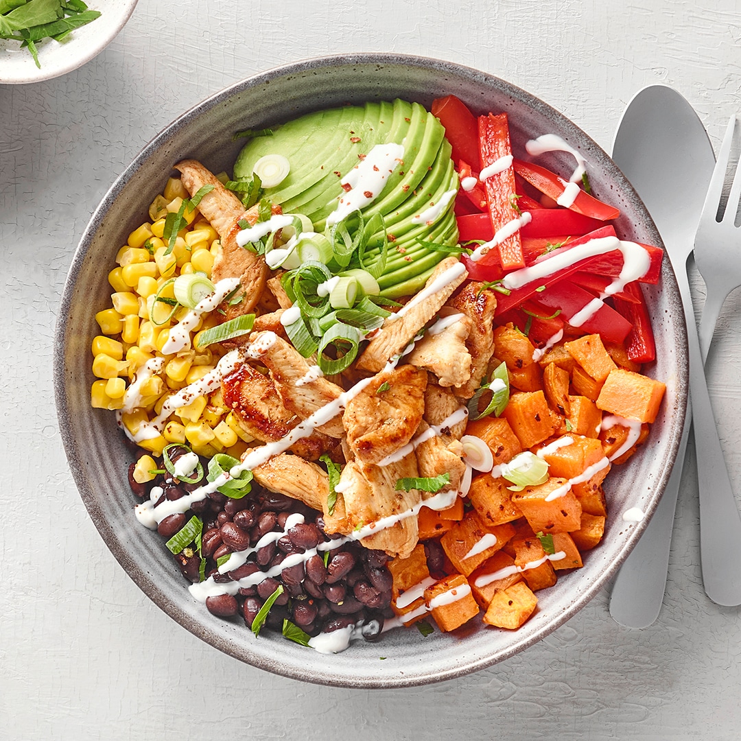 Bunte Bowl mit gegrilltem Hähnchen, Avocado, Mais, schwarzen Bohnen, Süßkartoffeln und Paprika, garniert mit Frühlingszwiebeln und Dressing.