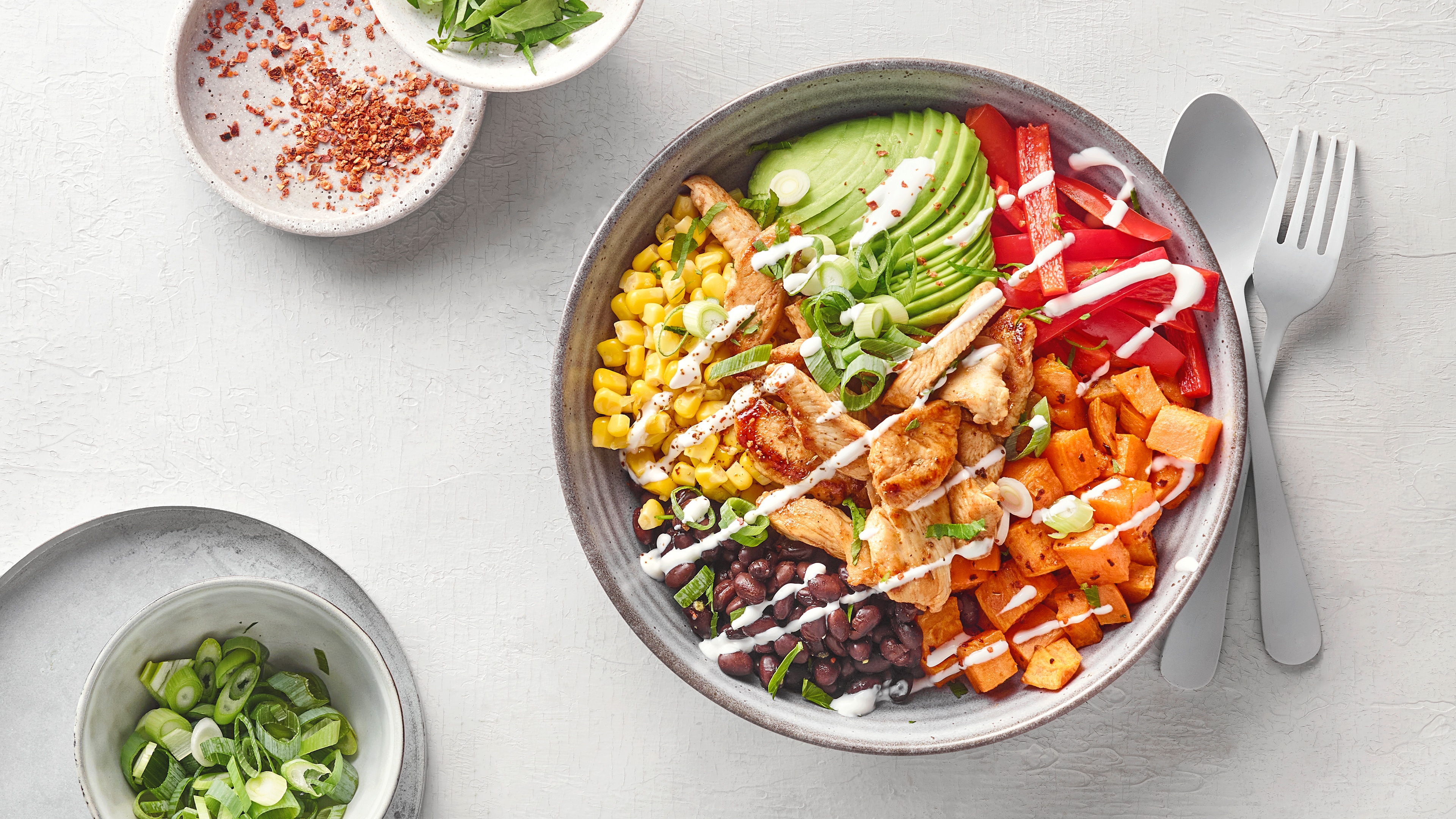 Bunte Bowl mit gegrilltem Hähnchen, Avocado, Mais, schwarzen Bohnen, Süßkartoffeln und Paprika, garniert mit Frühlingszwiebeln und Dressing.