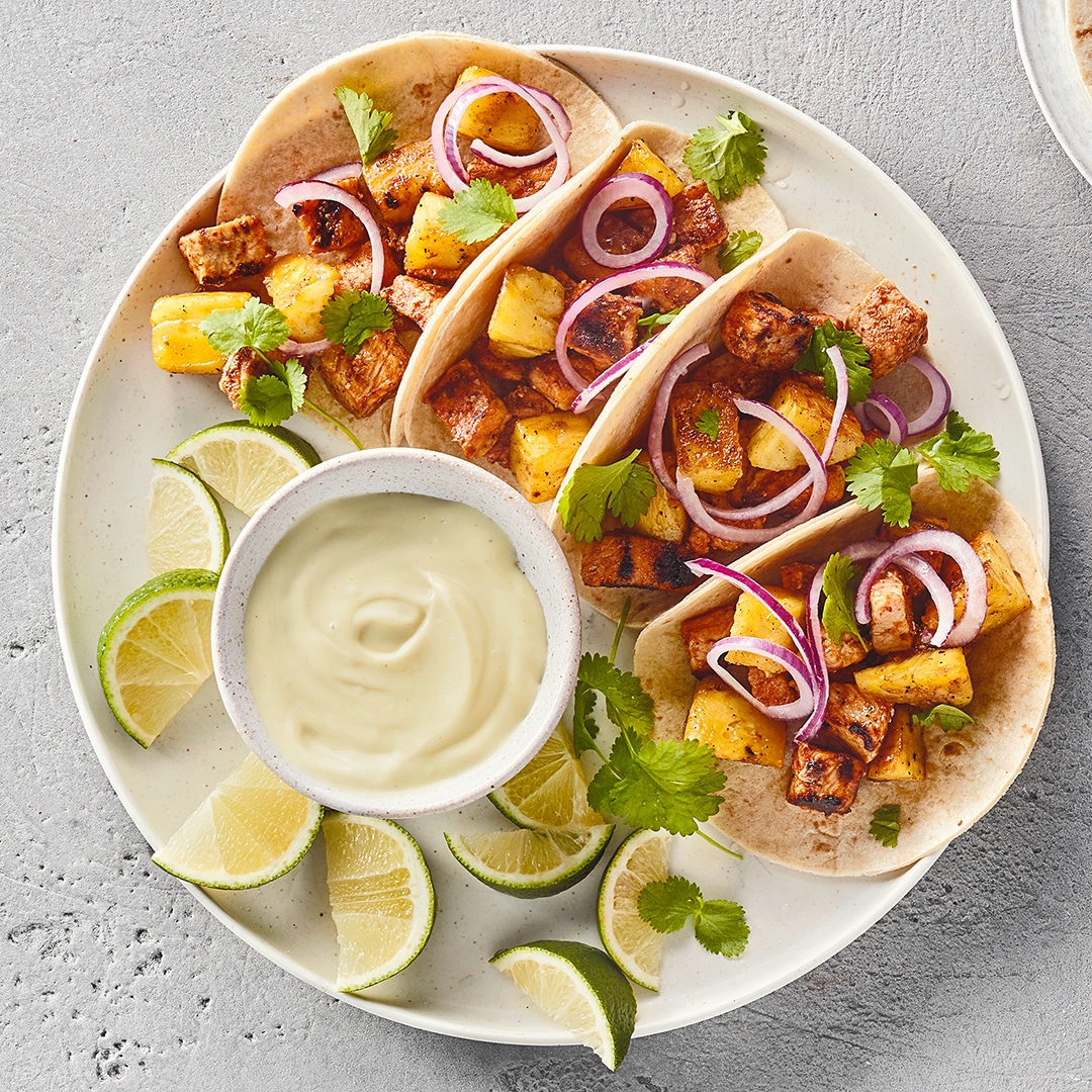 Ein Teller mit weichen Fladen, gefüllt mit Fleischwürfeln, roten Zwiebelringen & grünen Kräutern, daneben Limettenspalten