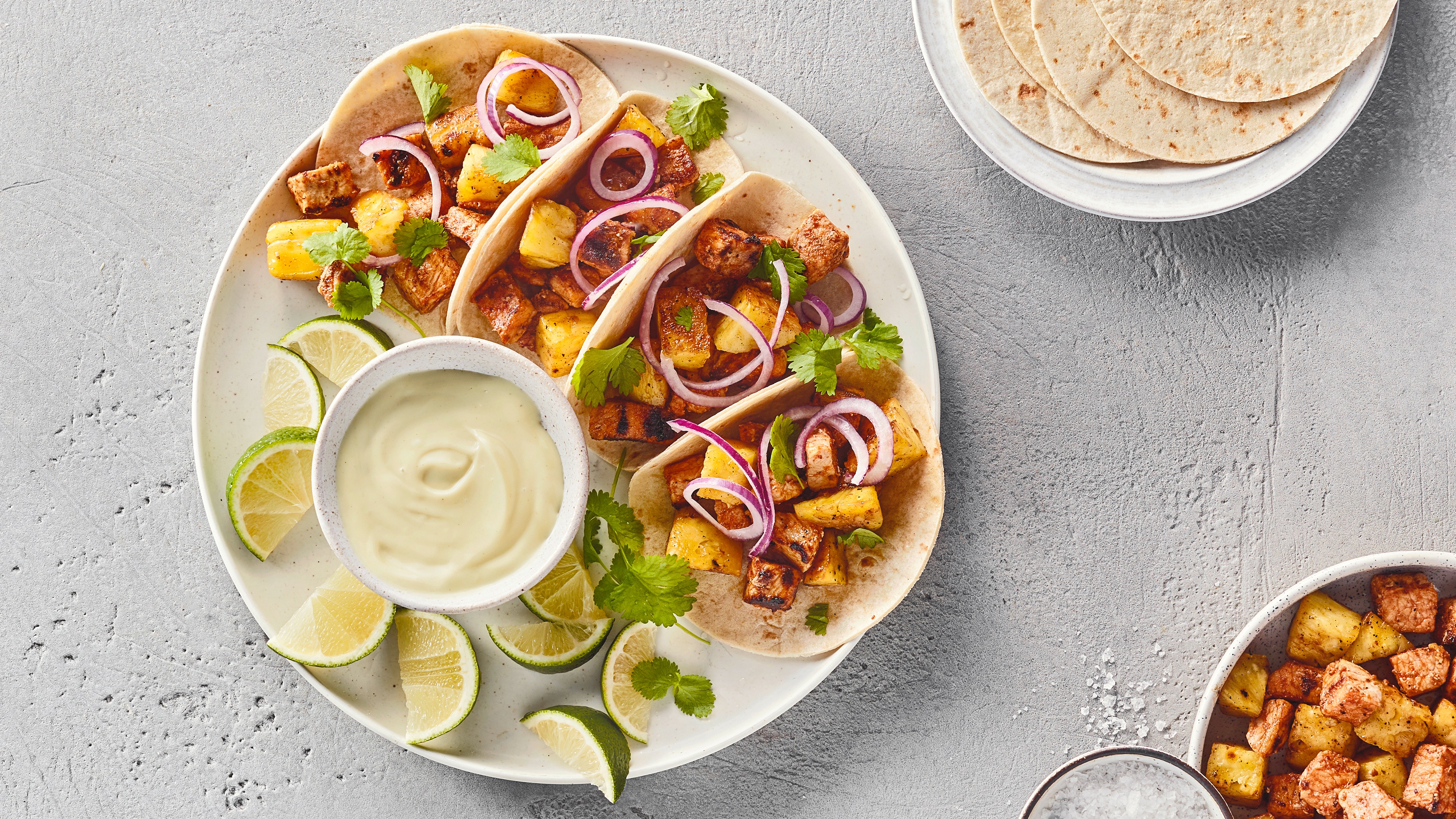 Ein Teller mit weichen Fladen, gefüllt mit Fleischwürfeln, roten Zwiebelringen & grünen Kräutern, daneben Limettenspalten