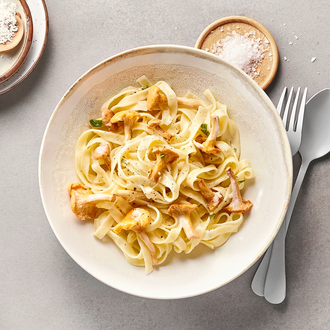 Tagliatelle mit Pfifferlingen in cremiger Sauce auf einem Teller, dazu geriebener Käse, Salz und Besteck auf hellem Untergrund.