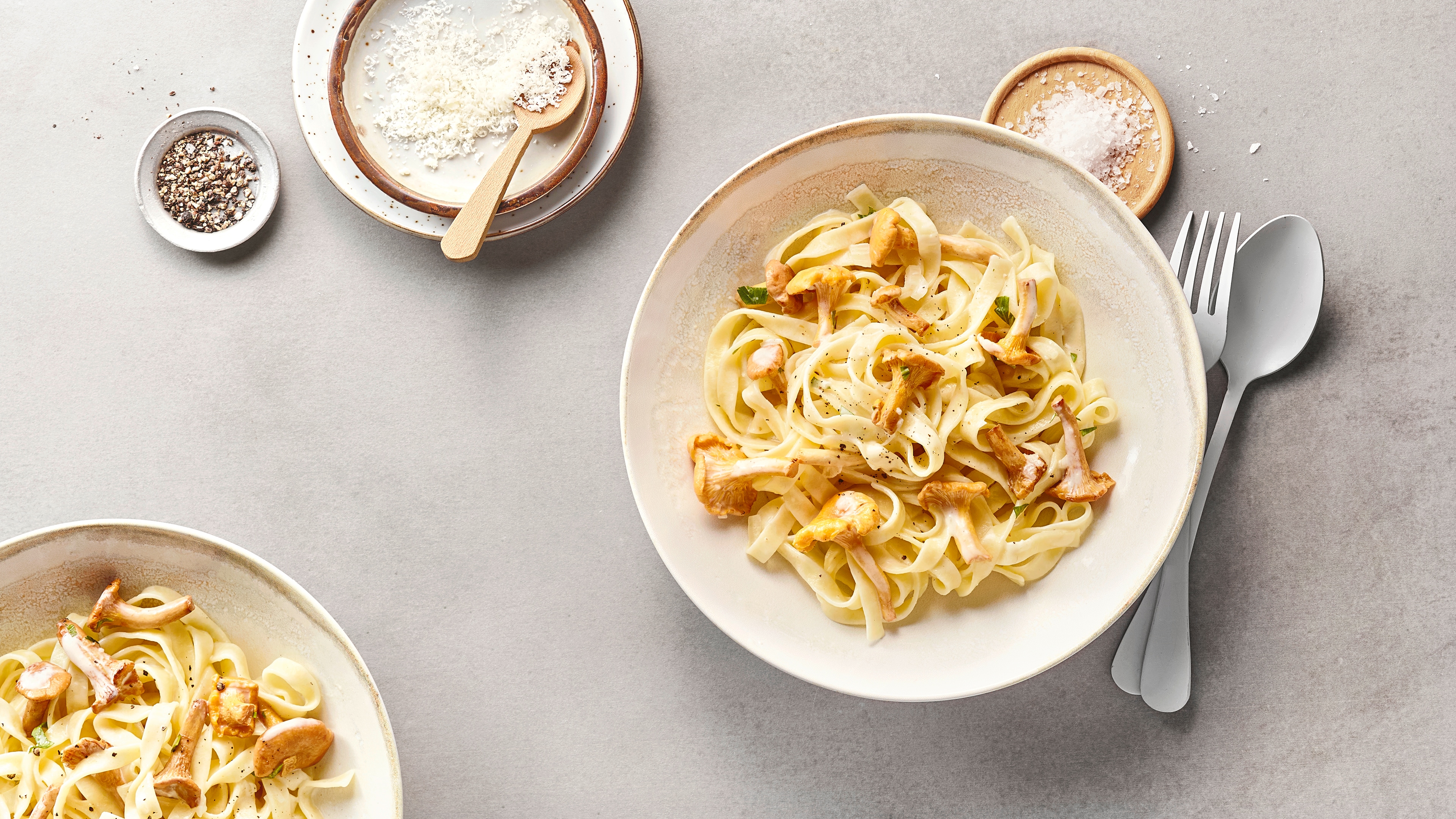 Tagliatelle mit Pfifferlingen in cremiger Sauce auf einem Teller, dazu geriebener Käse, Salz und Besteck auf hellem Untergrund.