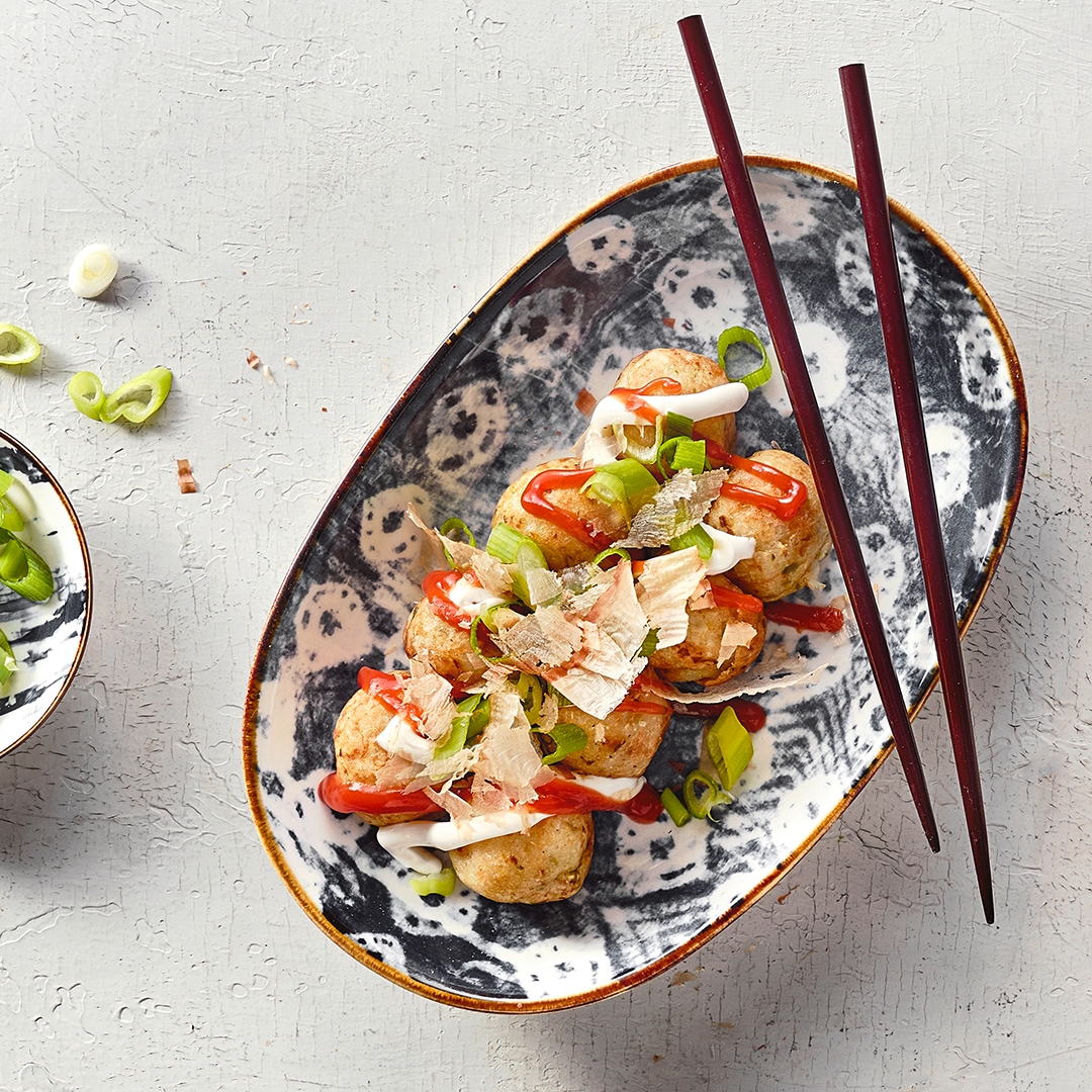 Ein Teller mit runden Bällchen, bedeckt mit roter und weißer Soße, Frühlingszwiebel und dünnen hellen Salatblättern.