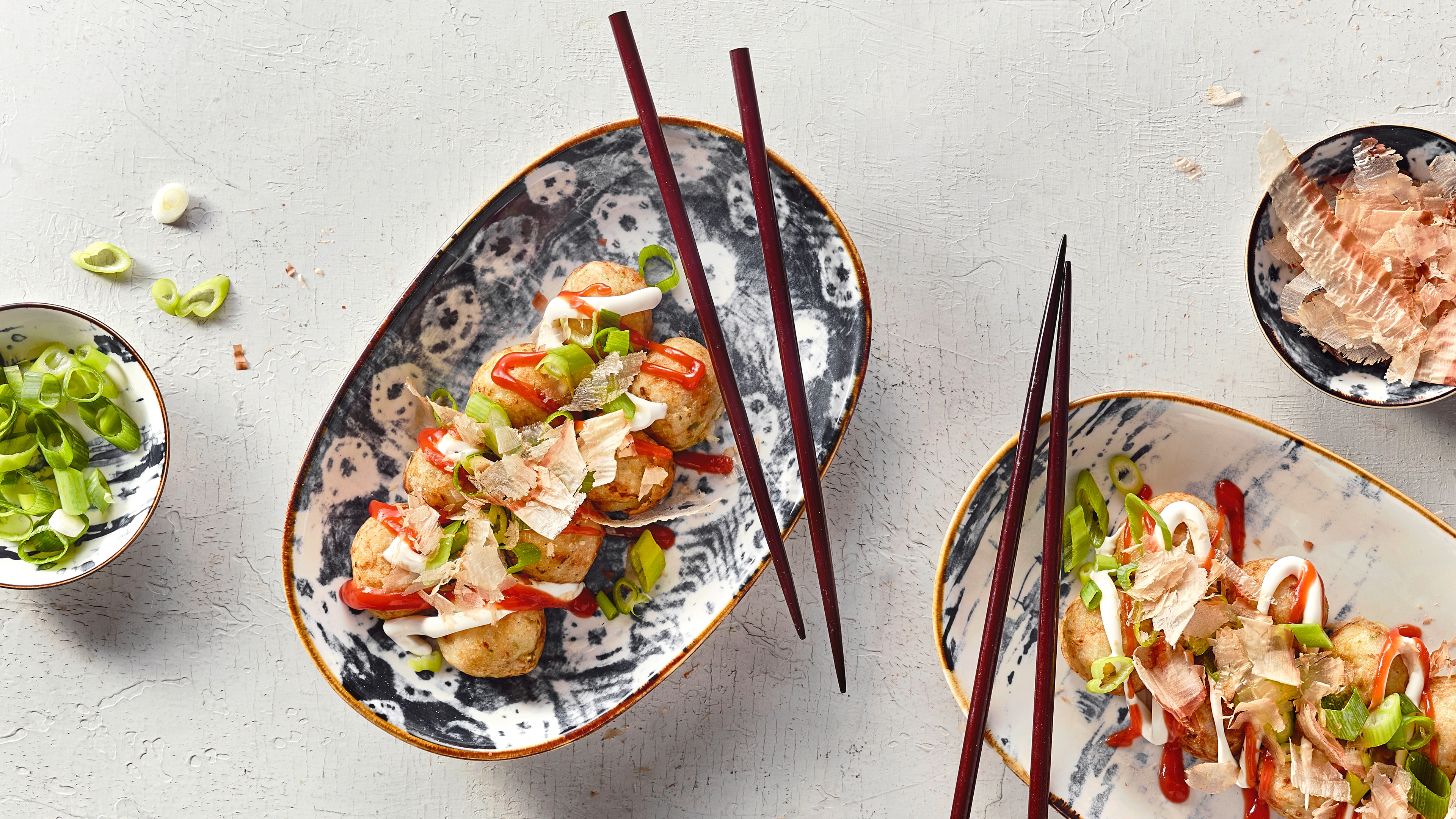 Ein Teller mit runden Bällchen, bedeckt mit roter und weißer Soße, Frühlingszwiebel und dünnen hellen Salatblättern.
