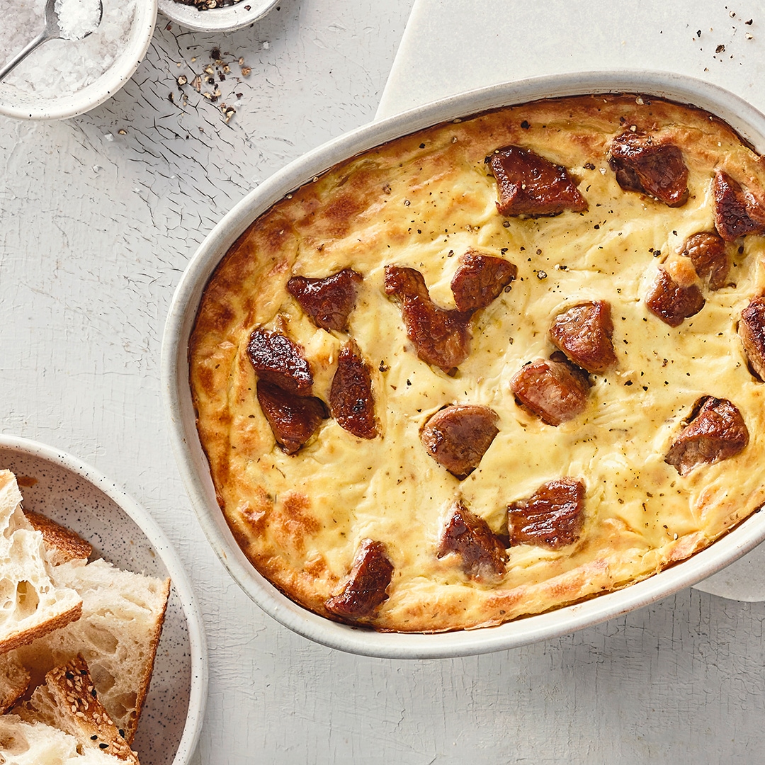 Eine Auflaufform mit goldbrauner, überbackener Masse, darin verteilt mehrere gebräunte Fleischstücke, daneben ein Teller mit hellen Brotscheiben.