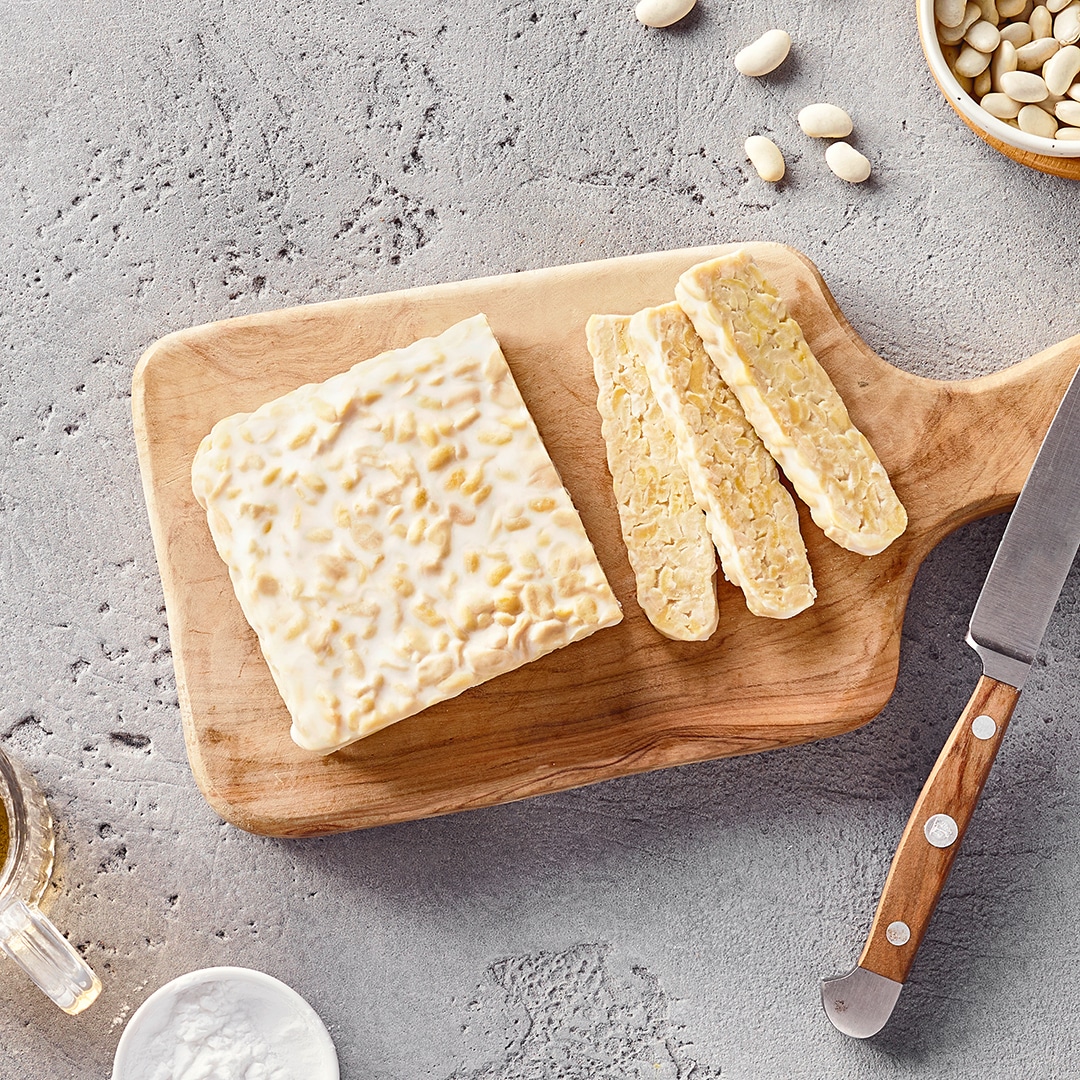Ein Brett mit weißem Tempeh, davon ein paar Scheiben abgeschnitten, daneben ein Schälchen mit weißen Bohnen