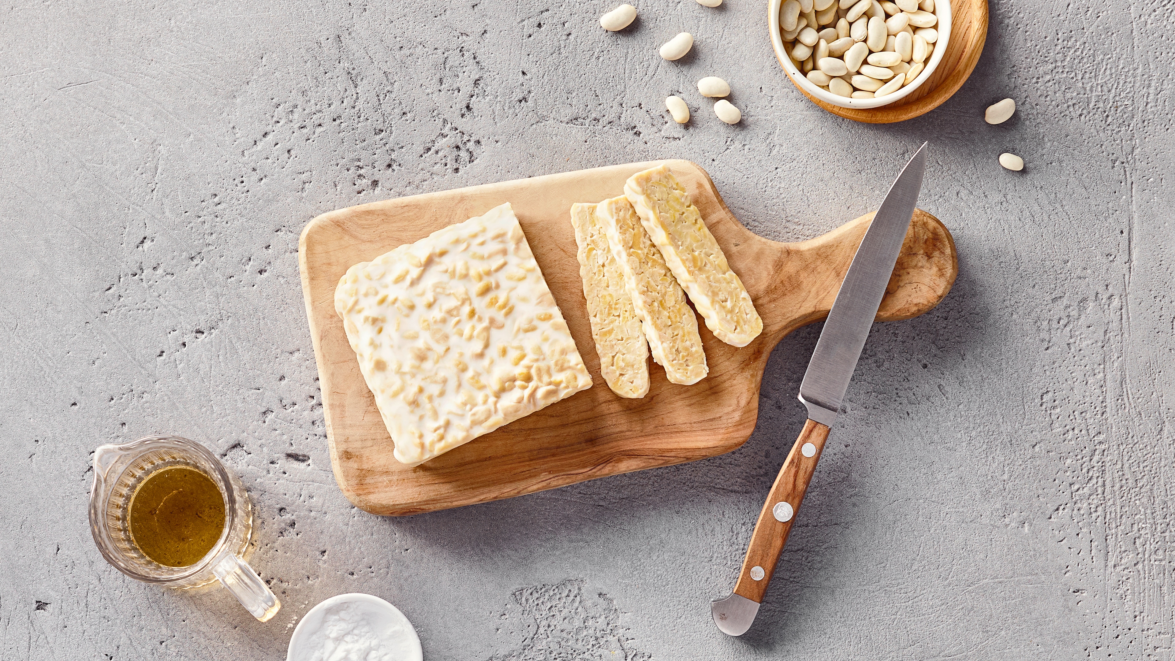 Ein Brett mit weißem Tempeh, davon ein paar Scheiben abgeschnitten, daneben ein Schälchen mit weißen Bohnen