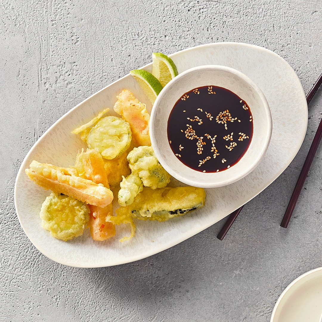 Ein Teller mit hellgrünen, knusprigen Gemüsestücken im Teigmantel, zwei Limettenstücken und einer Schale dunkler Soße mit Sesam.