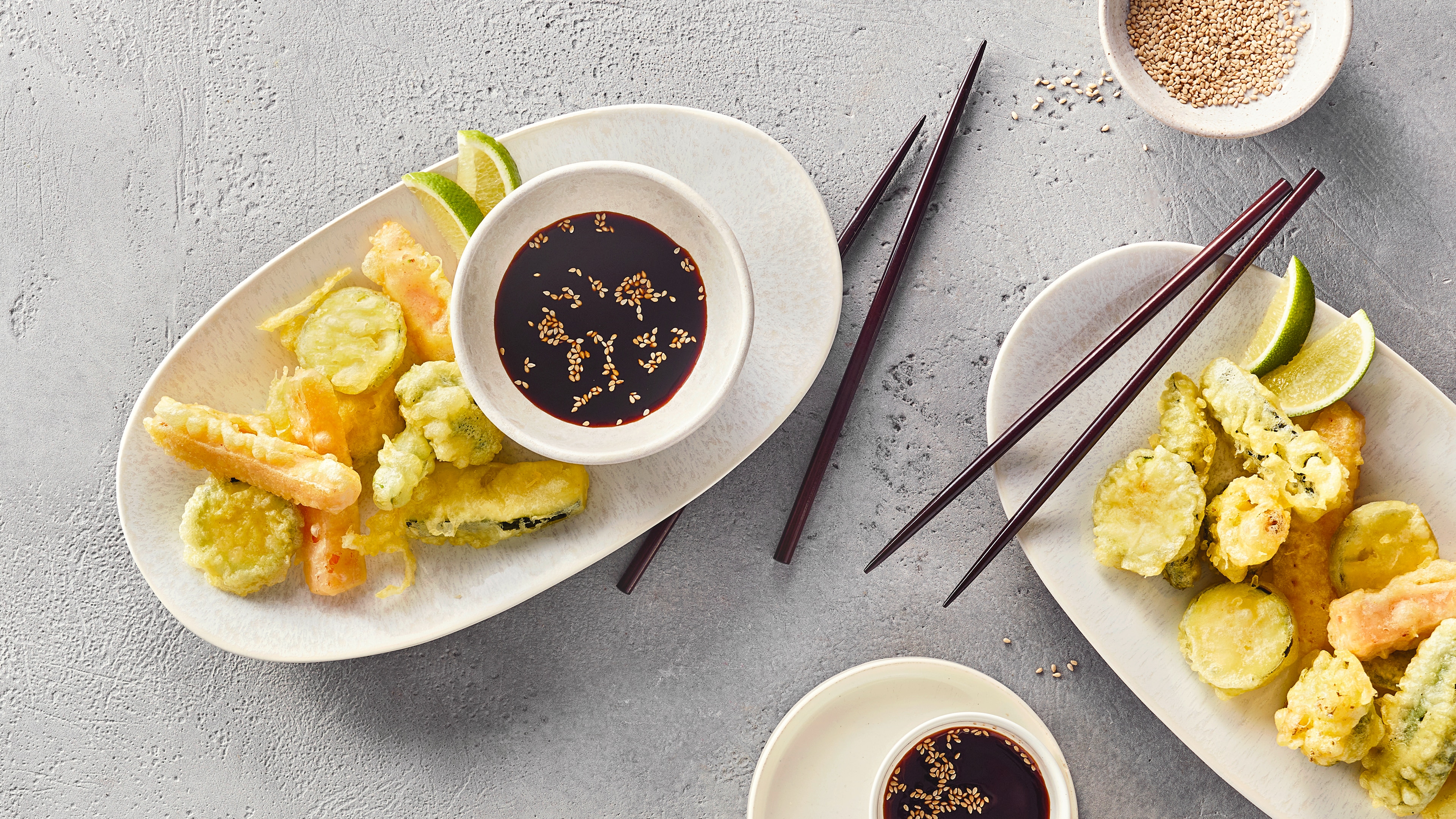 Ein Teller mit hellgrünen, knusprigen Gemüsestücken im Teigmantel, zwei Limettenstücken und einer Schale dunkler Soße mit Sesam.