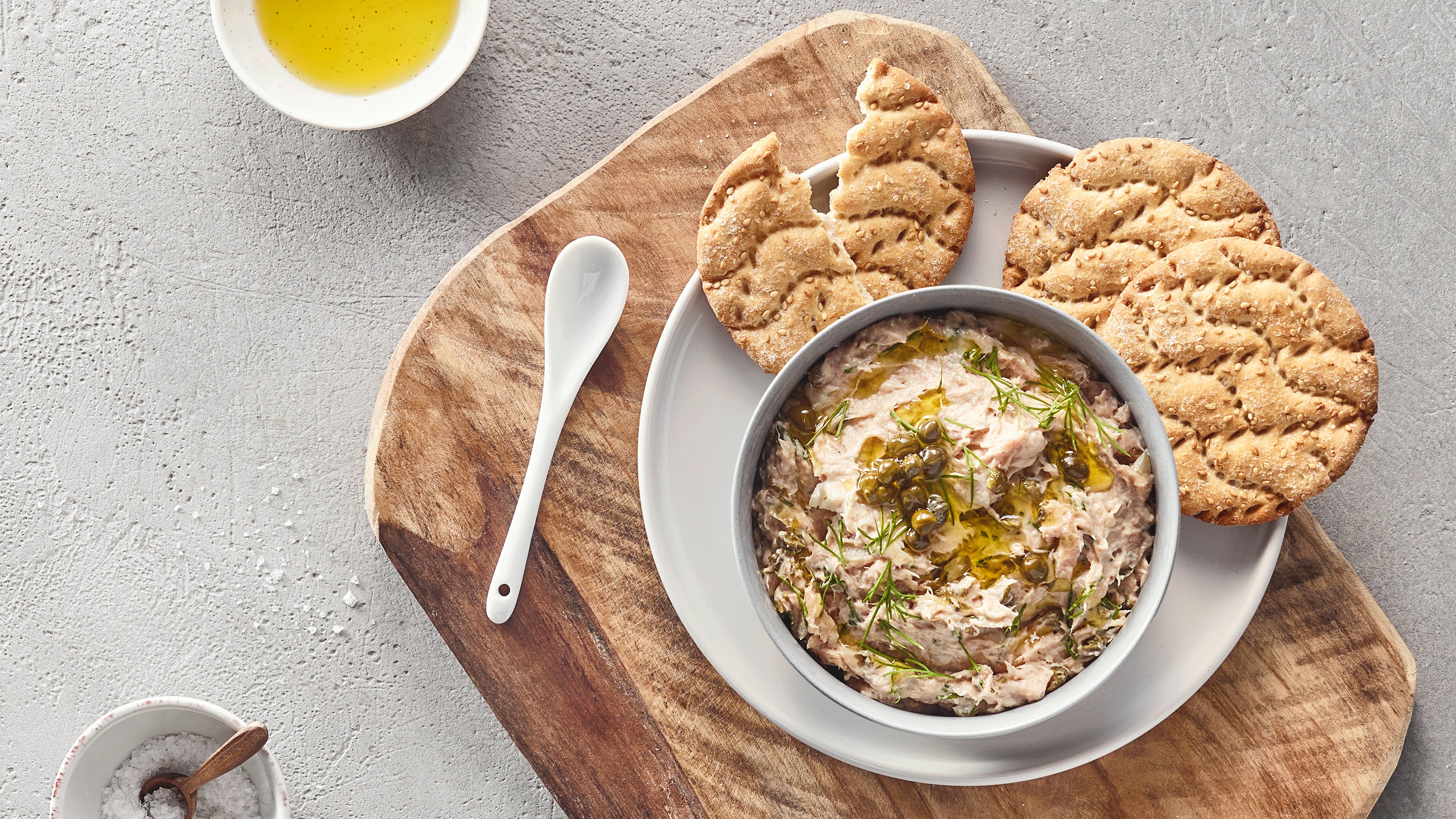 Schale mit cremigem Aufstrich, der mit Kräutern, Öl und Kapern garniert ist. Daneben runde Cracker.