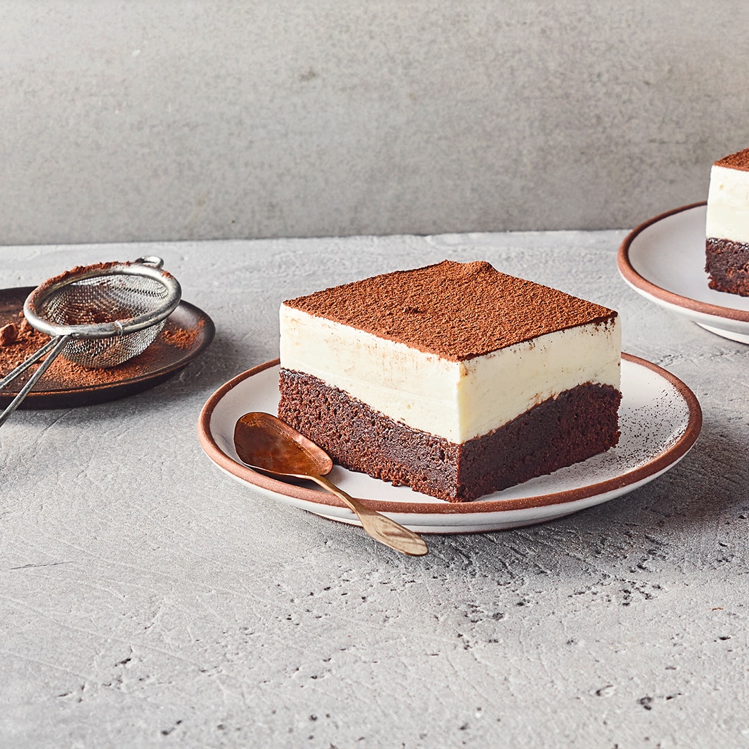 Ein Teller mit einem Stück Schichtdessert aus Schokolade und Creme, bestreut mit Kakaopulver, daneben liegt ein Löffel