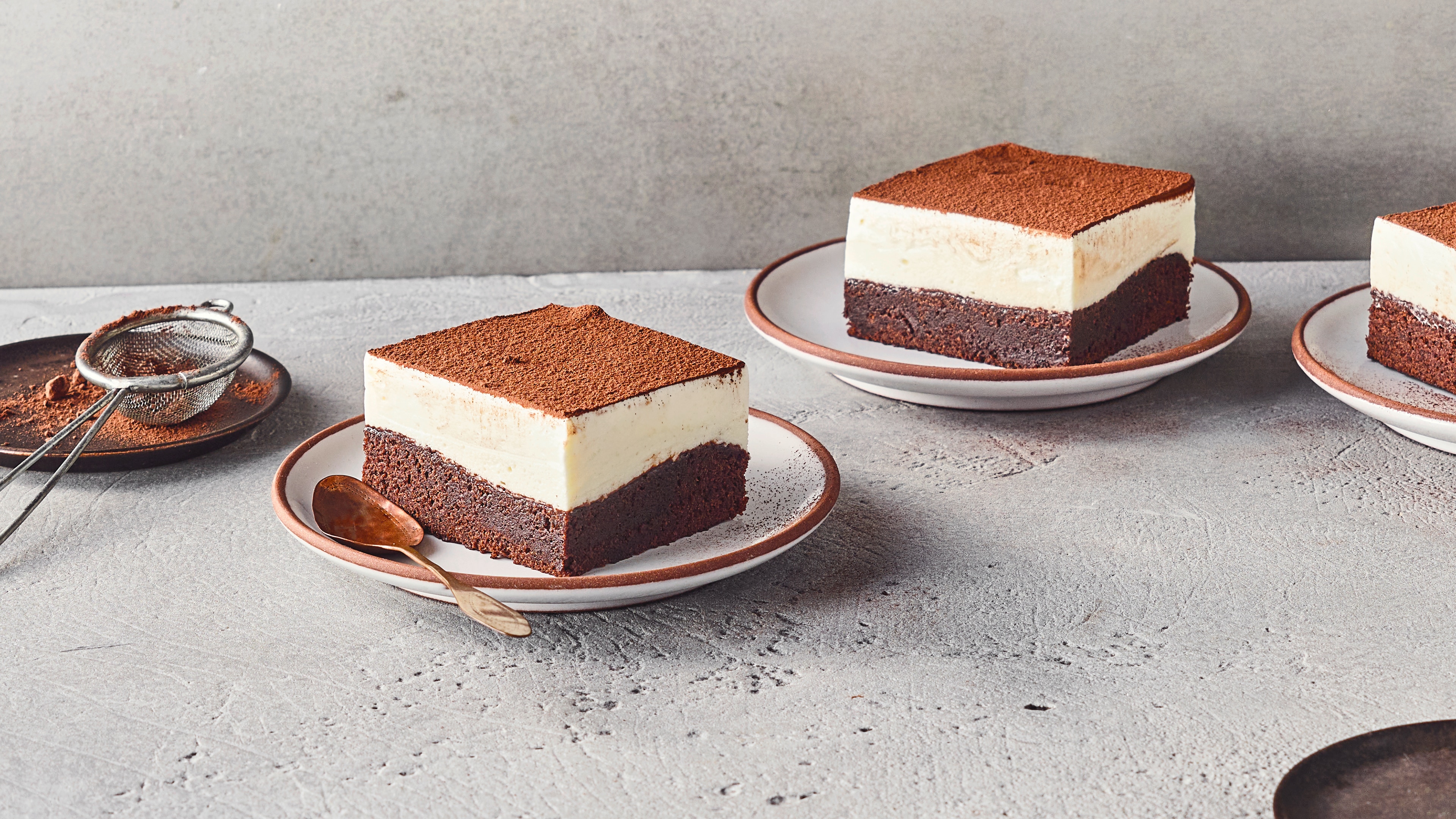 Ein Teller mit einem Stück Schichtdessert aus Schokolade und Creme, bestreut mit Kakaopulver, daneben liegt ein Löffel