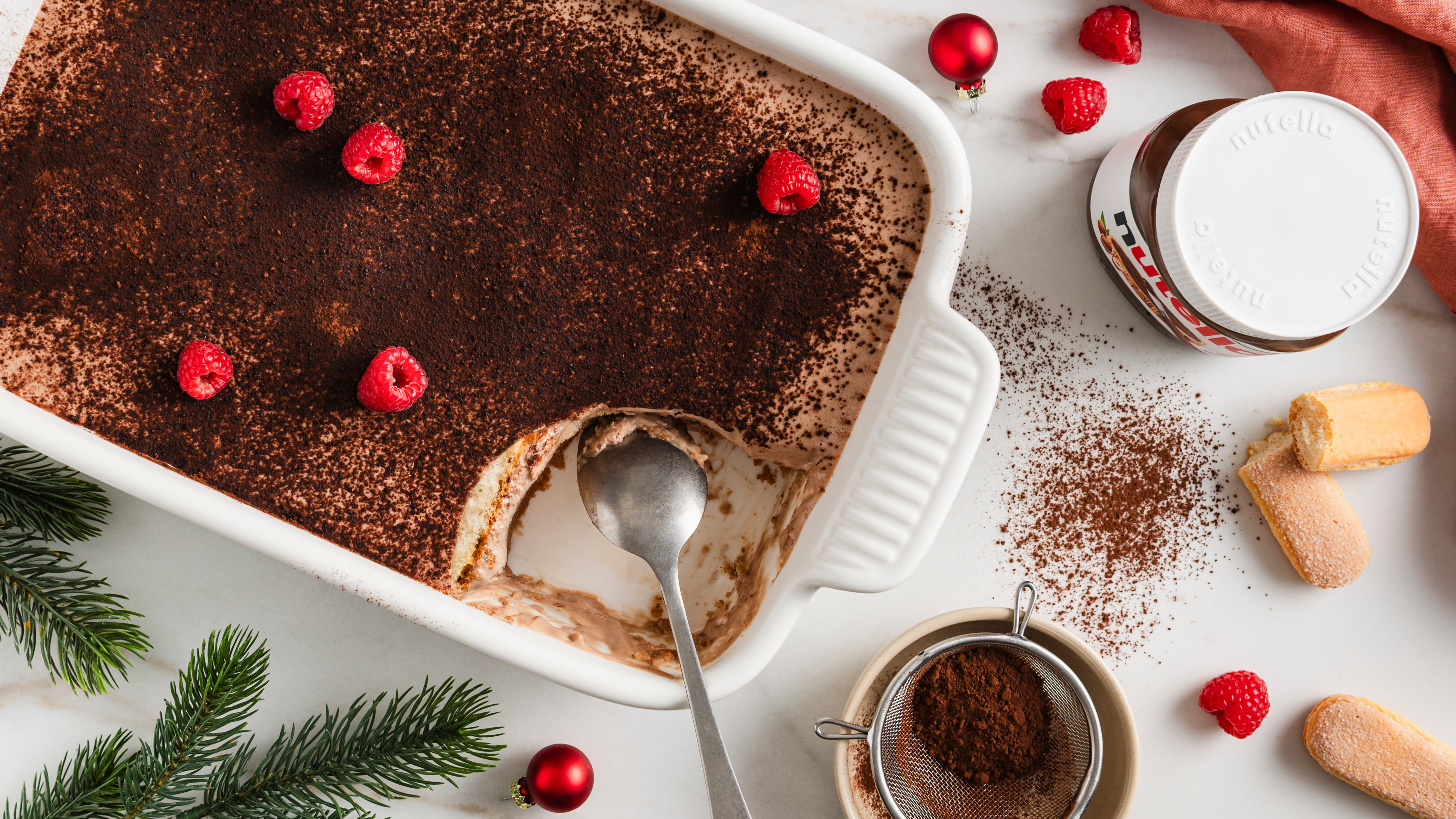 Eine Auflaufform mit cremigem Dessert, bestäubt mit dunklem Kakaopulver, teils ausgelöffelt, garniert mit frischen Himbeeren