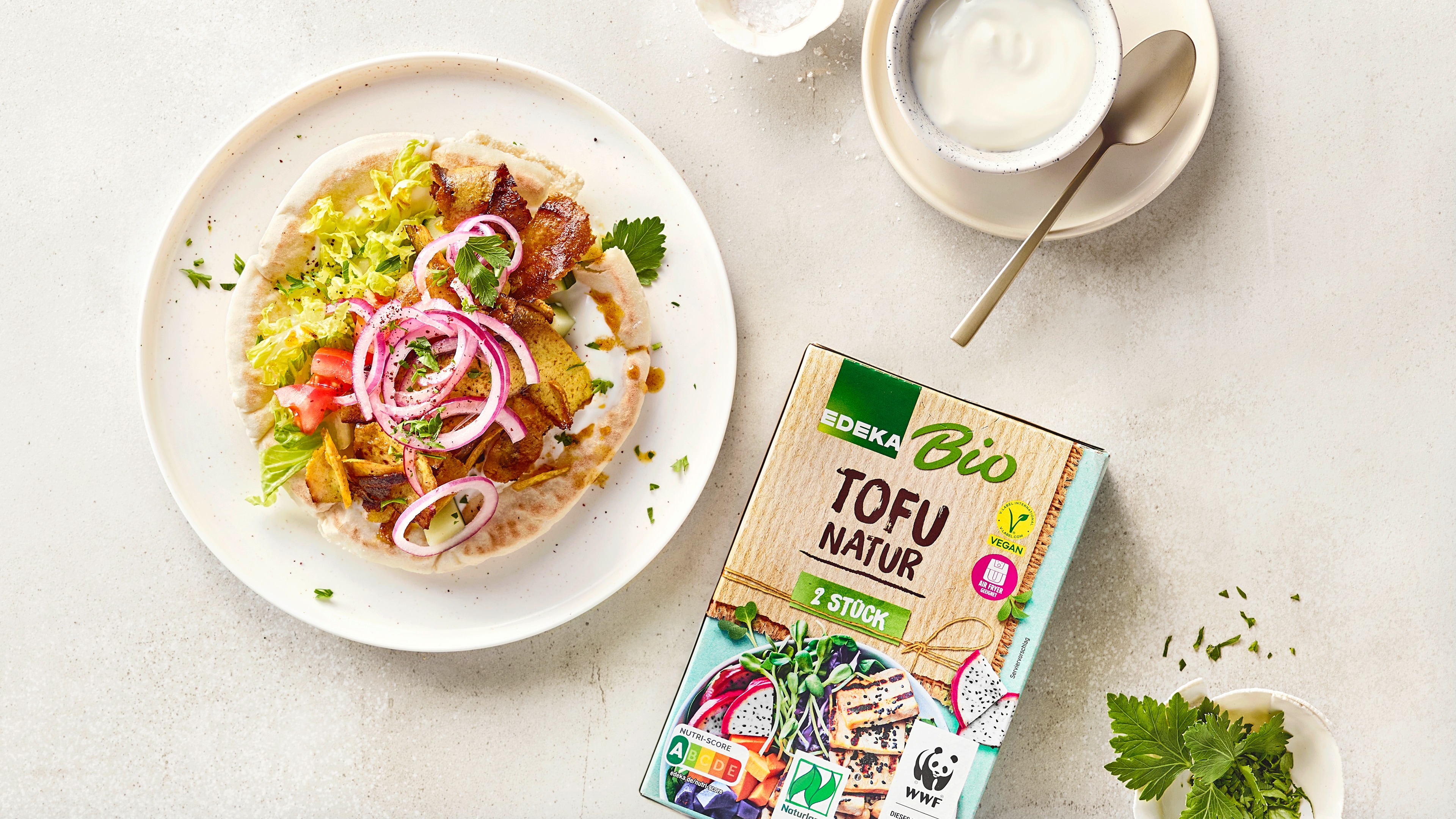 Gefülltes Fladenbrot mit Salat, roten Zwiebeln, Gemüse und gebratenem Tofu auf weißem Teller, serviert mit Joghurt-Dip; daneben EDEKA Bio Tofu Natur.