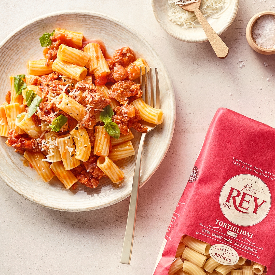 Teller mit Tortiglioni in Tomaten-Hackfleischsauce, garniert mit geriebenem Käse und Basilikum, daneben eine Schale mit Käse und Salz sowie Pasta Rey Tortiglioni Verpackung.