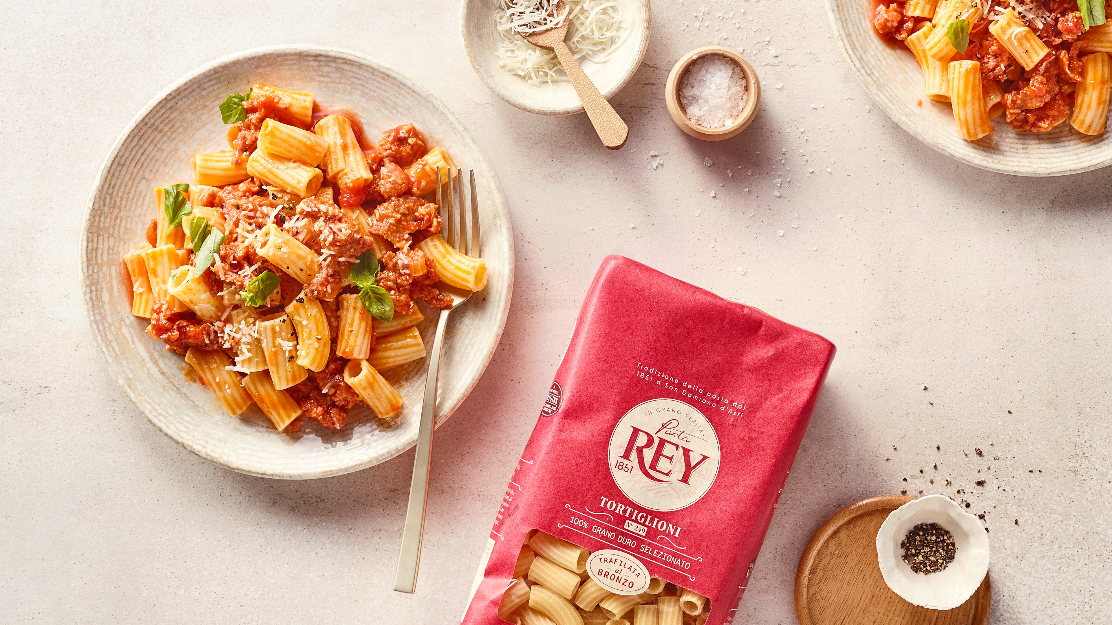 Zwei Teller mit Tortiglioni in Tomaten-Hackfleischsauce, garniert mit geriebenem Käse und Basilikum; daneben Schalen mit Käse und Salz sowie Pasta Rey Tortiglioni Verpackung.