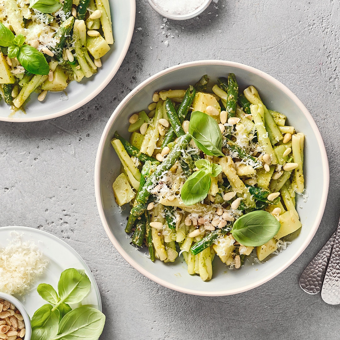 Eine Schüssel mit grünen und gelben Bohnen, Kartoffelstücken, grünem Pesto, Pinienkernen & Käseraspeln mit Basilikumblättern.
