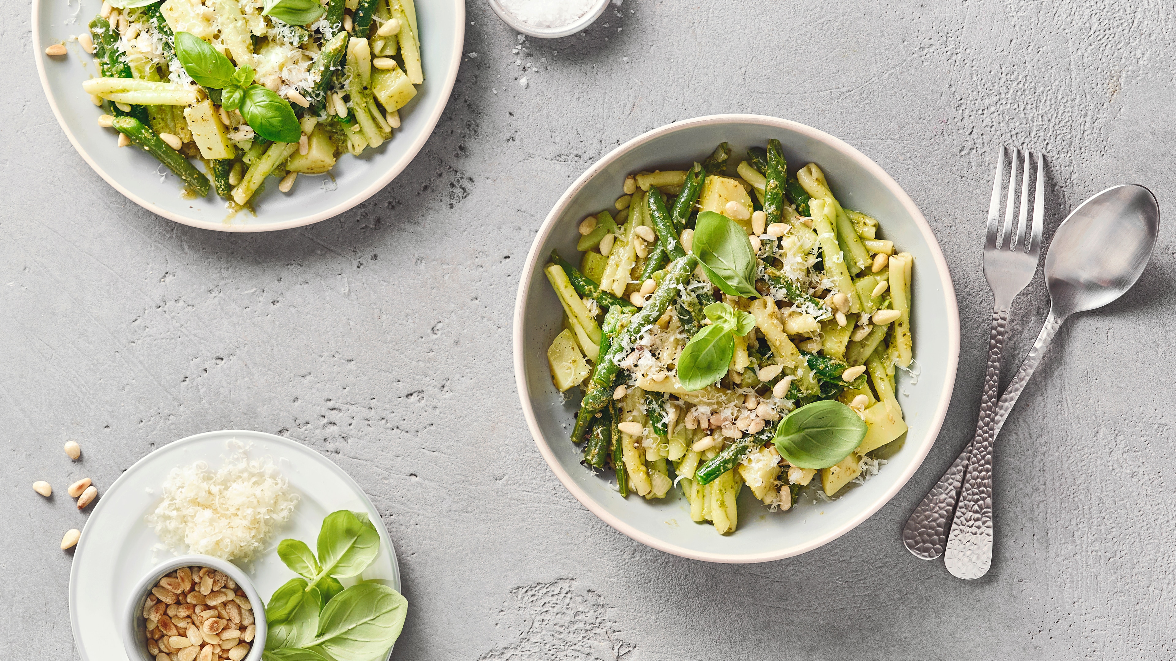 Eine Schüssel mit grünen und gelben Bohnen, Kartoffelstücken, grünem Pesto, Pinienkernen & Käseraspeln mit Basilikumblättern.