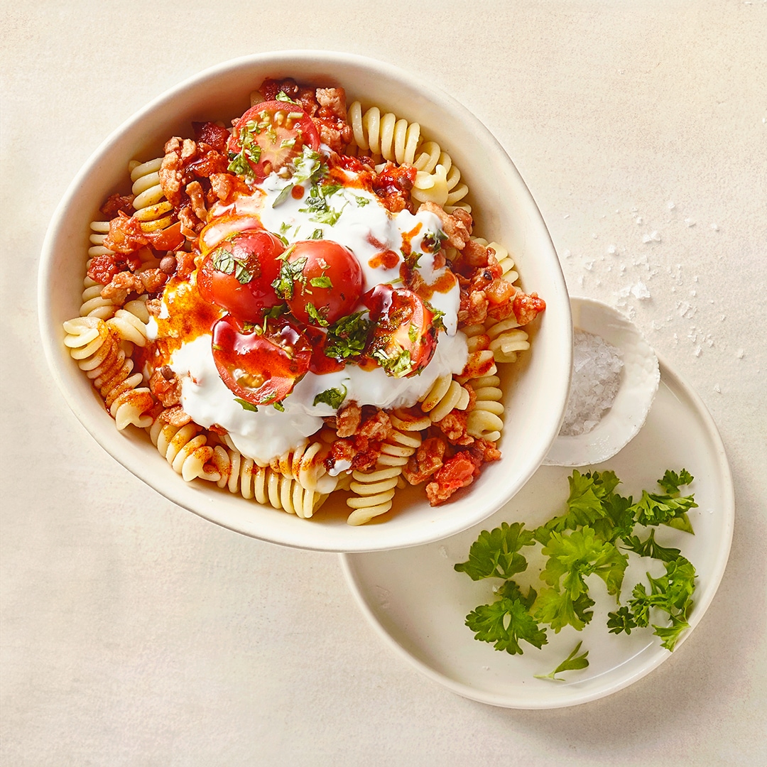 Eine Schale mit Spiralnudeln, kräftiger Tomaten-Hacksoße, garniert mit saftigen Tomatenhälften, weißem Joghurt und grünen Kräutern