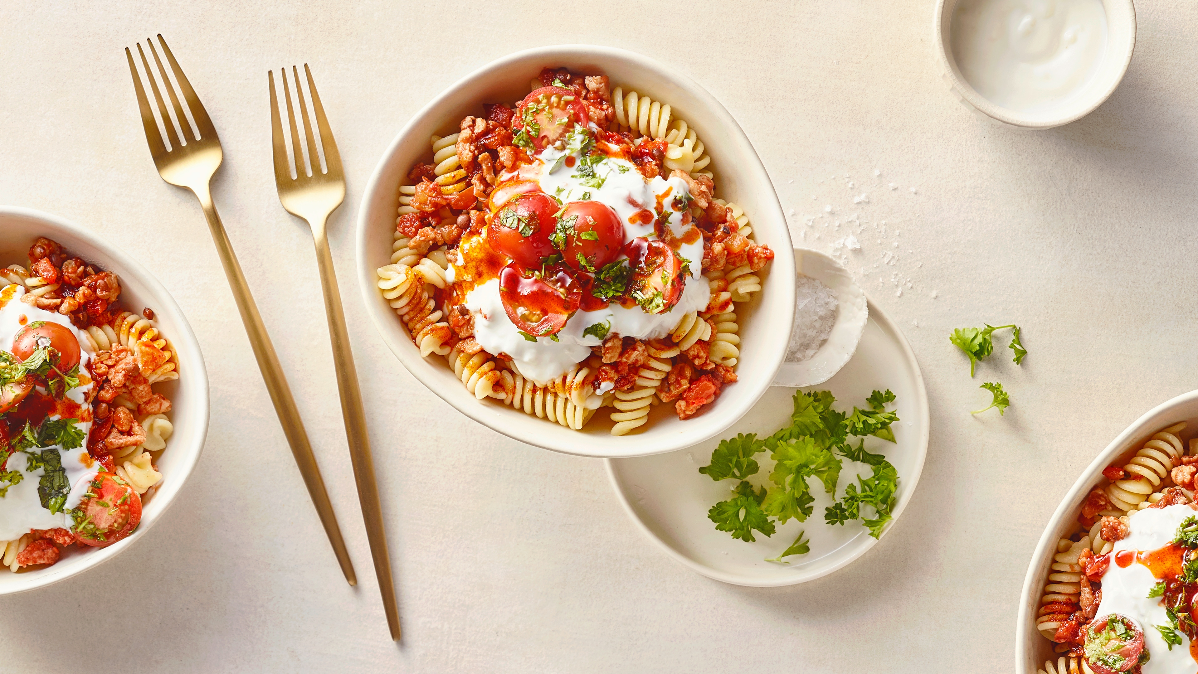 Eine Schale mit Spiralnudeln, kräftiger Tomaten-Hacksoße, garniert mit saftigen Tomatenhälften, weißem Joghurt und grünen Kräutern