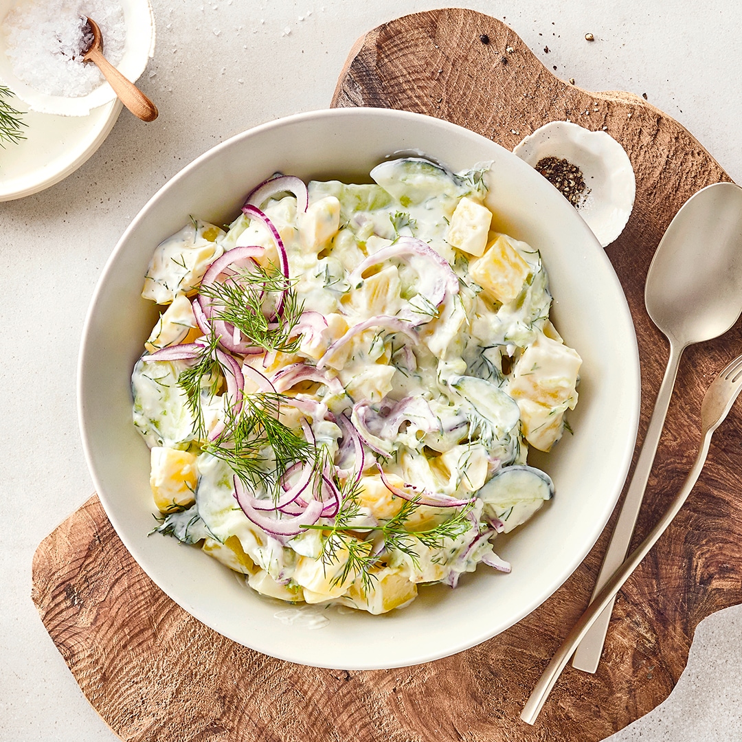 Kartoffel-Gurken-Salat mit roten Zwiebeln und frischem Dill in cremigem Dressing, serviert in einer Schüssel auf einem Holzbrett mit Kräutern und Gewürzen.