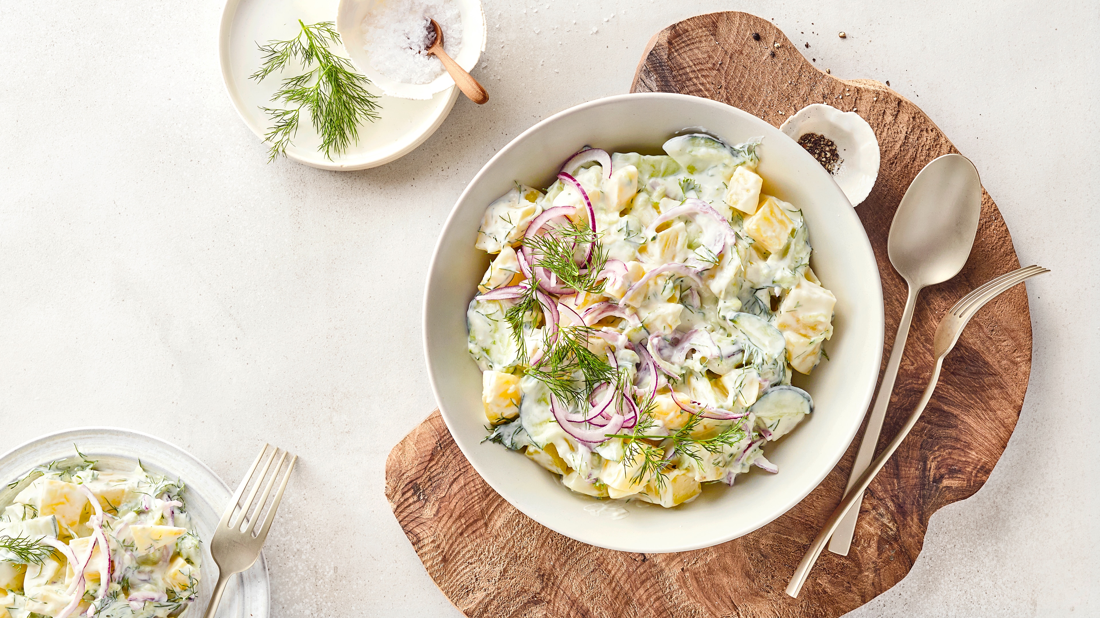 Kartoffel-Gurken-Salat mit roten Zwiebeln und frischem Dill in cremigem Dressing, serviert in einer Schüssel auf einem Holzbrett mit Kräutern und Gewürzen.