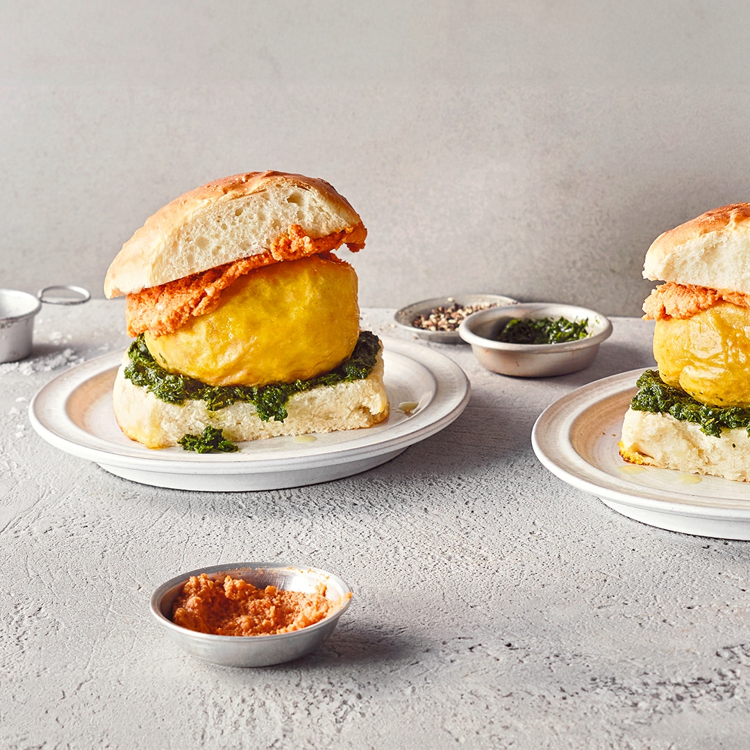 Ein Teller mit hellem Brötchen, gelbem Ball, grüner Kräuterpaste und bräunlich-roter Paste auf dem oberen Brötchen.