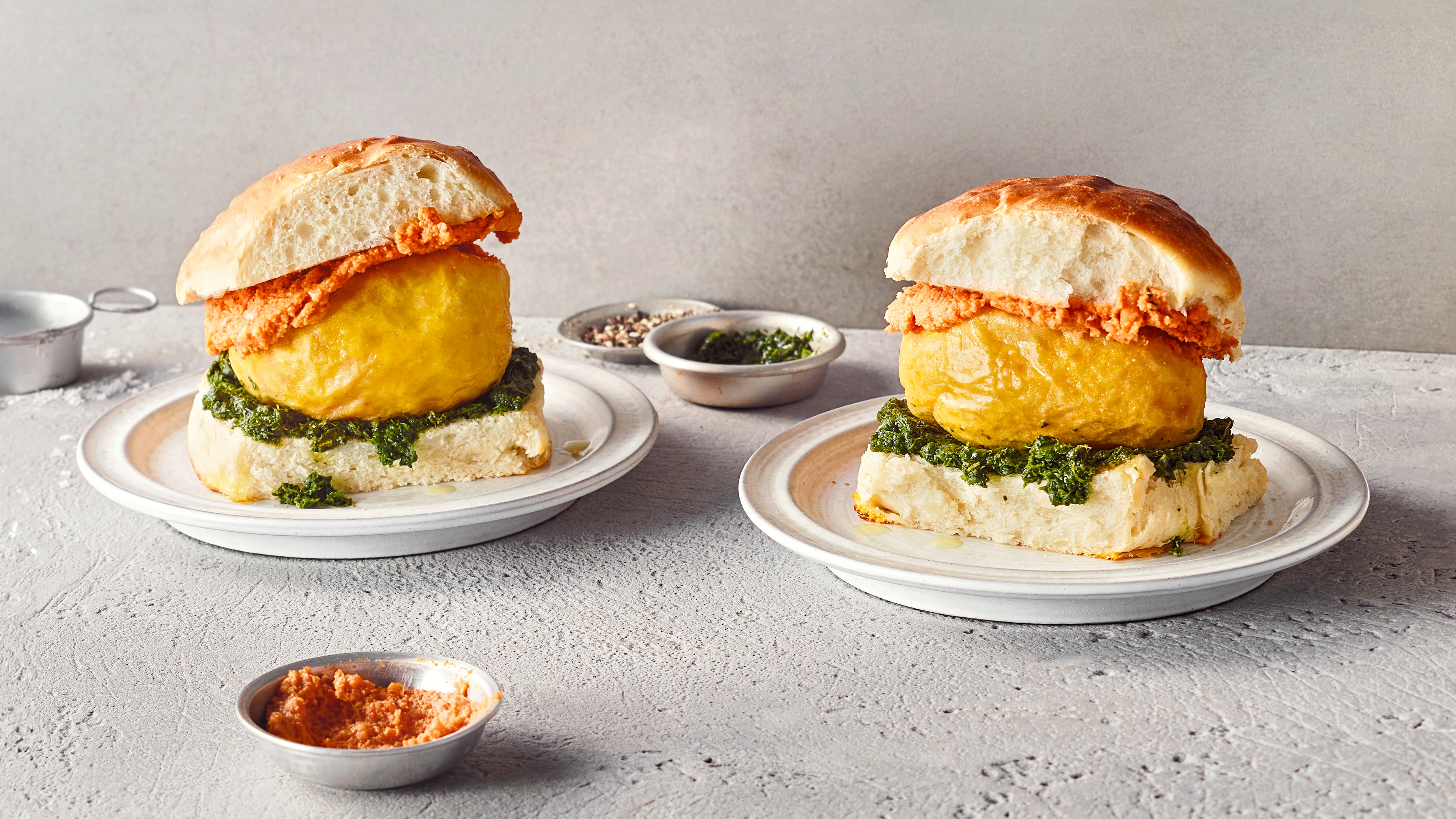 Ein Teller mit hellem Brötchen, gelbem Ball, grüner Kräuterpaste und bräunlich-roter Paste auf dem oberen Brötchen.
