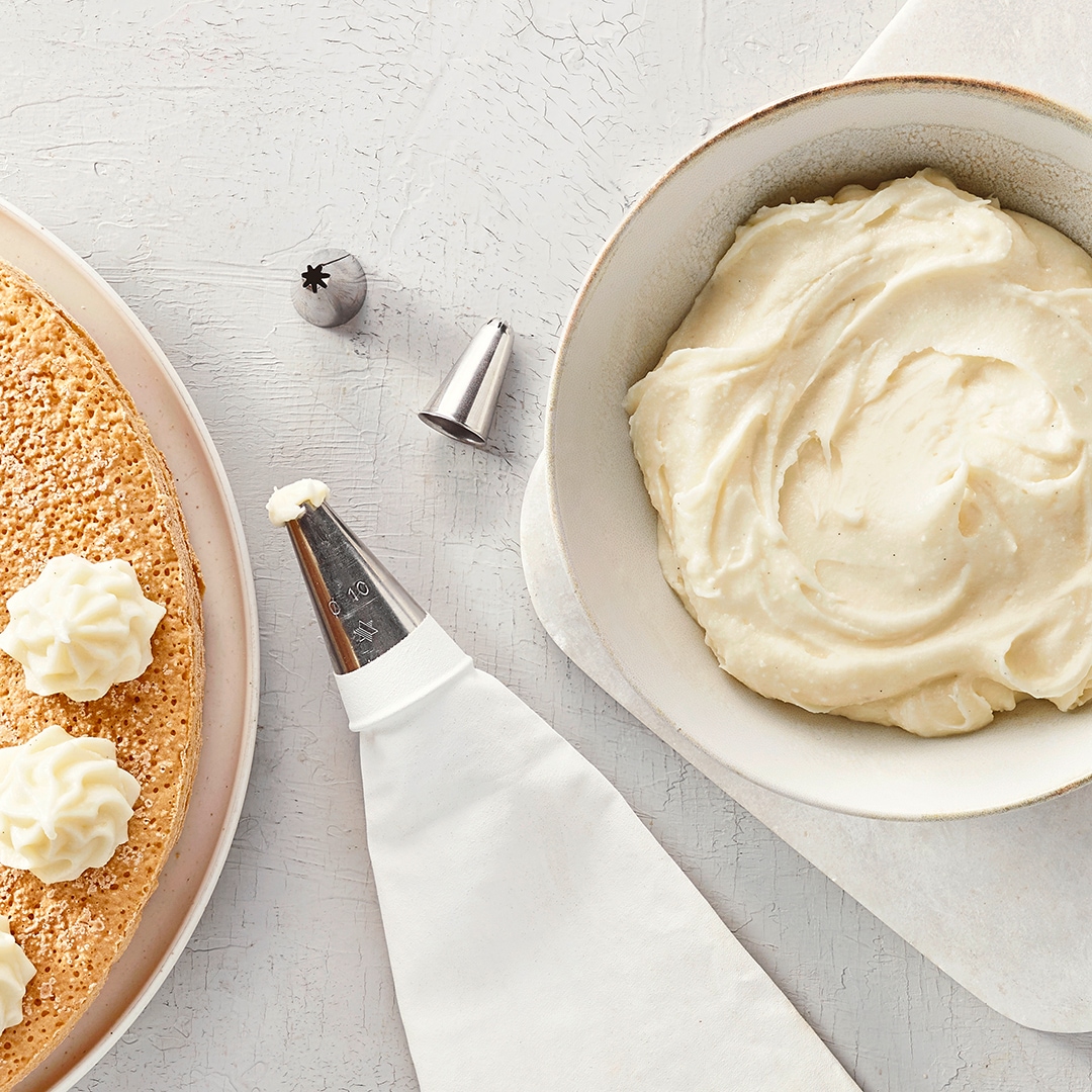 Eine Schüssel mit heller, aufgeschlagener Creme, daneben ein Spritzbeutel und eine heller Tortenboden